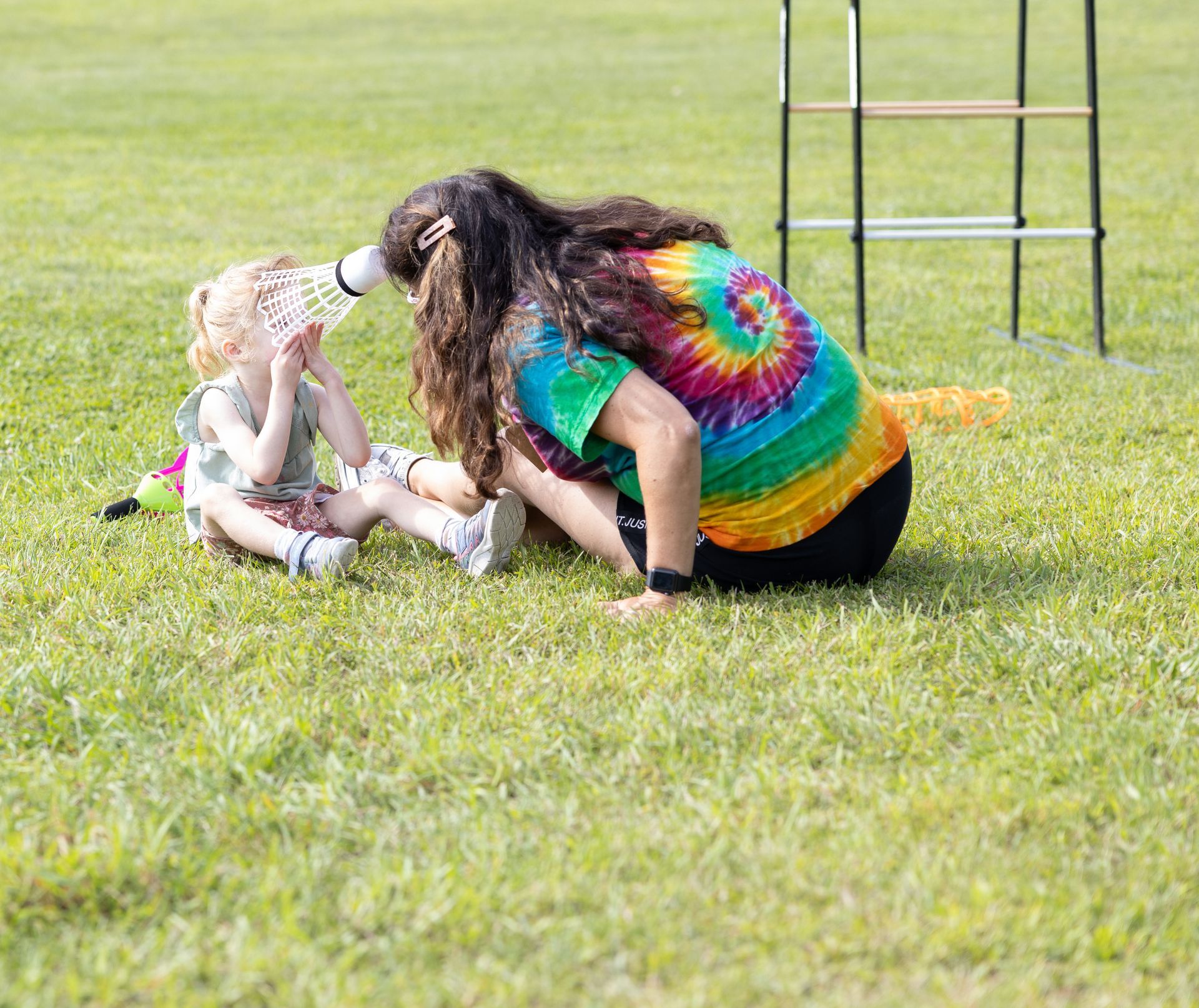 Person with tie-dye shirt and child on grass playing with a badminton shuttlecock near a net.