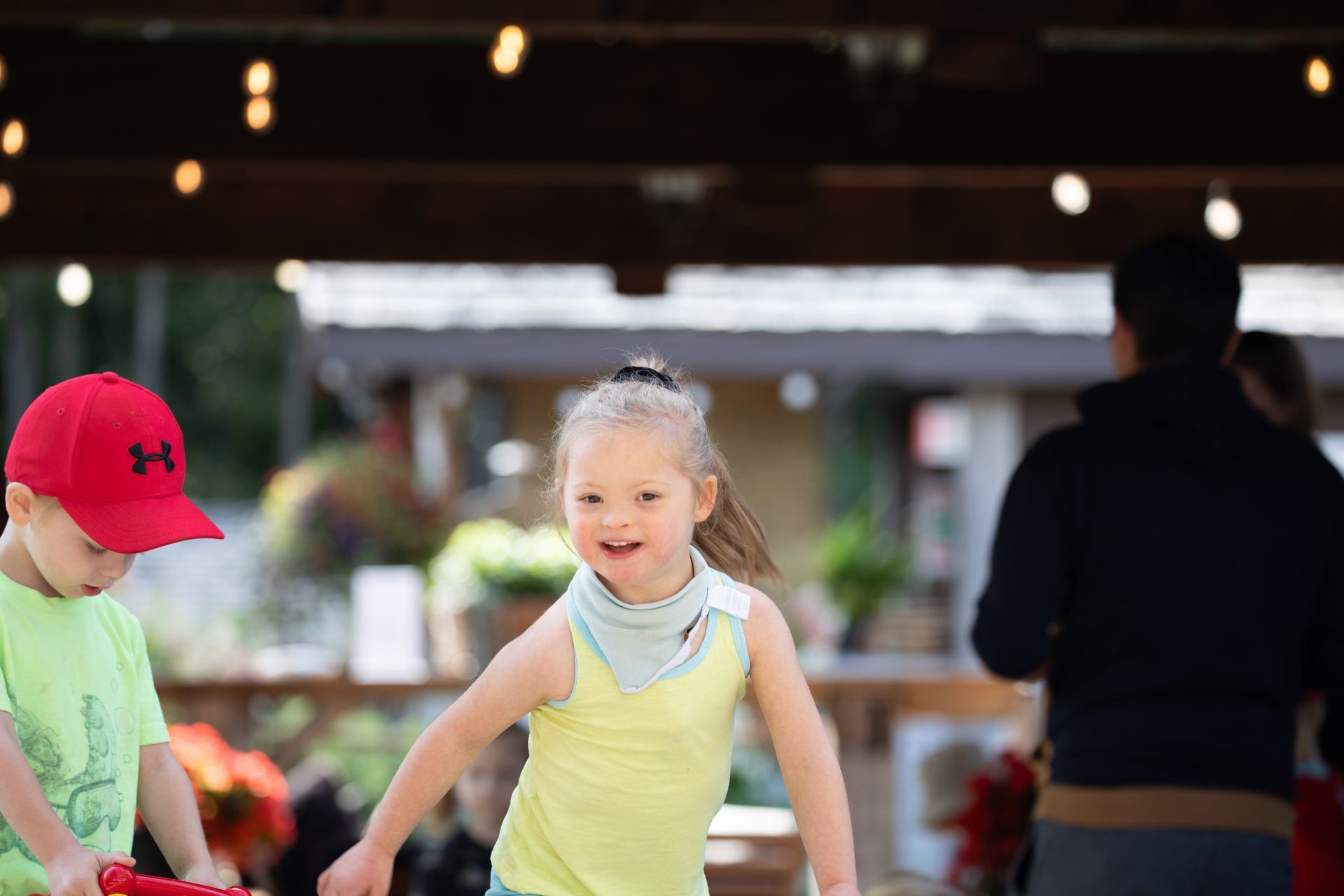 Girl smiles, runs forward at outdoor event, near boy in red cap and other people.