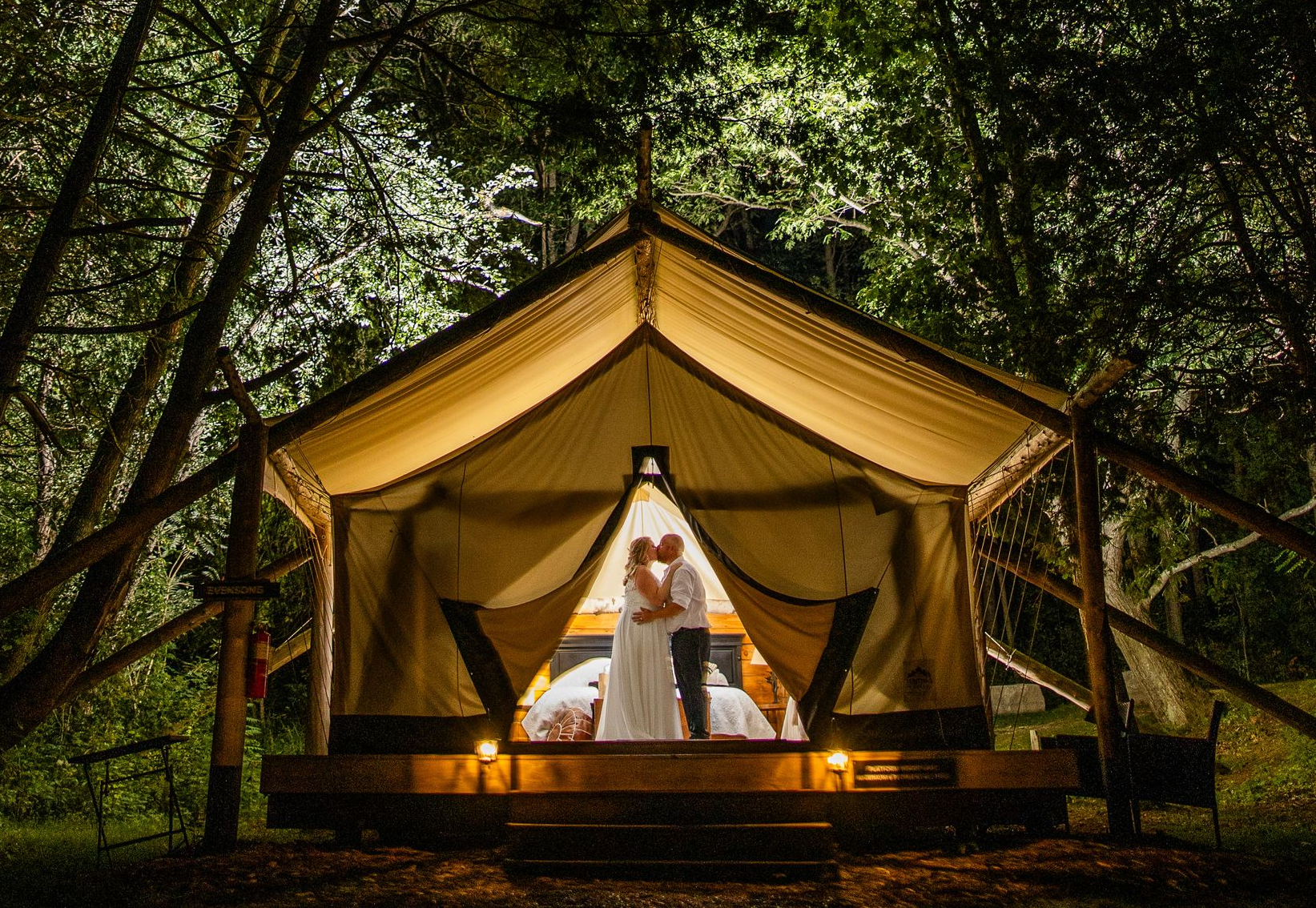 Couple embracing in a lit glamping tent in a dark forest. Warm light emanates from within.