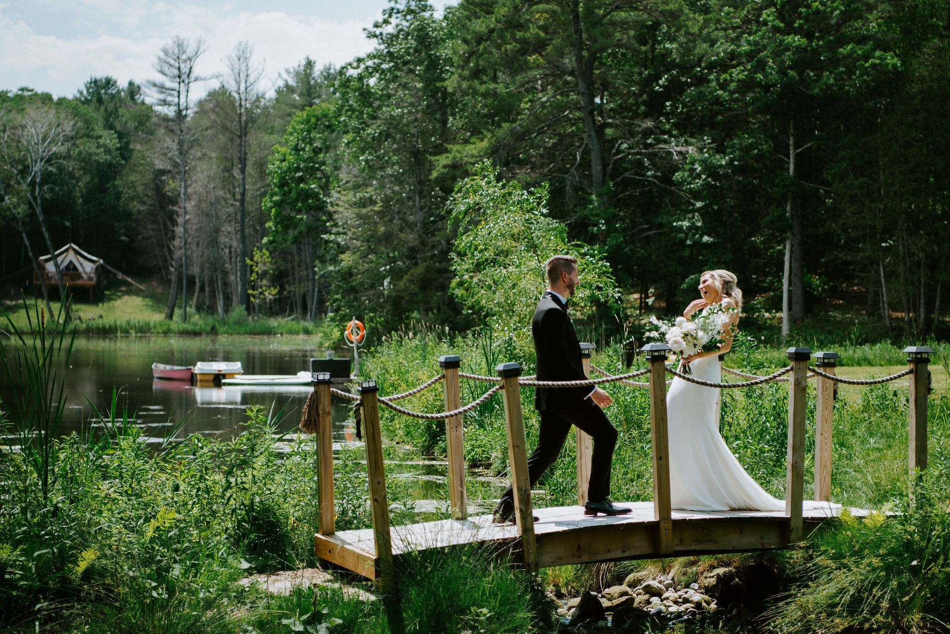 Bride and groom on a wooden bridge, looking at each other near a lake and trees.