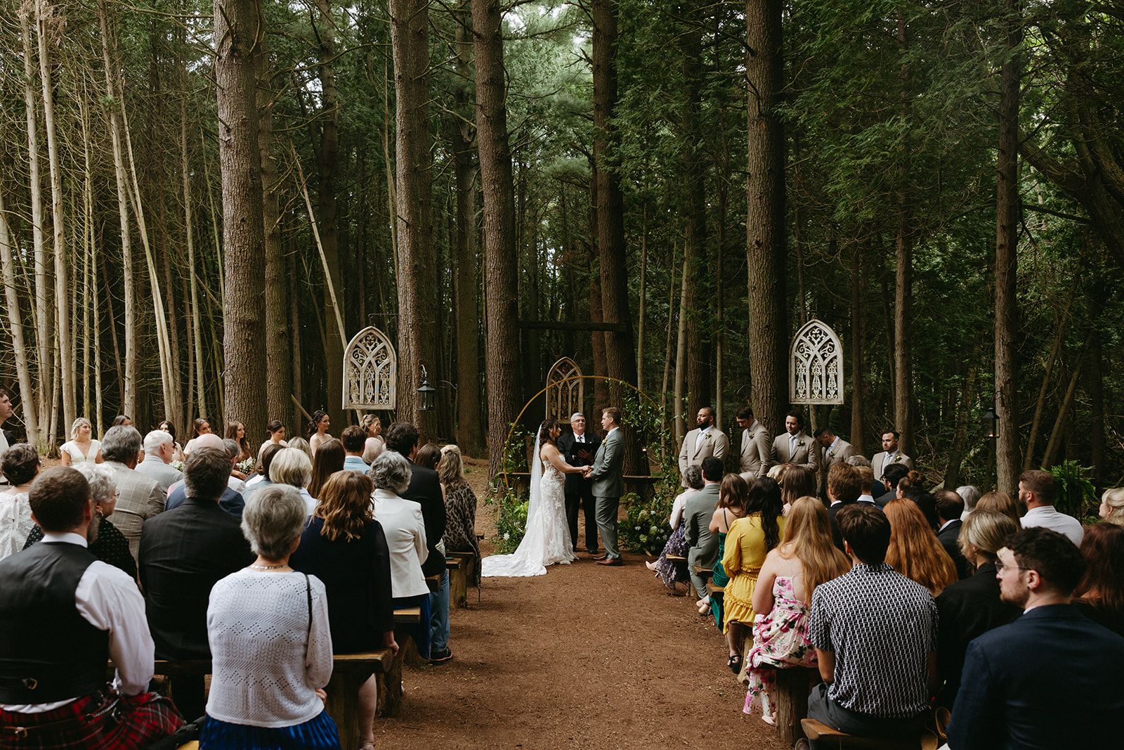 Wedding ceremony in a forest, bride and groom at altar, guests seated on benches.