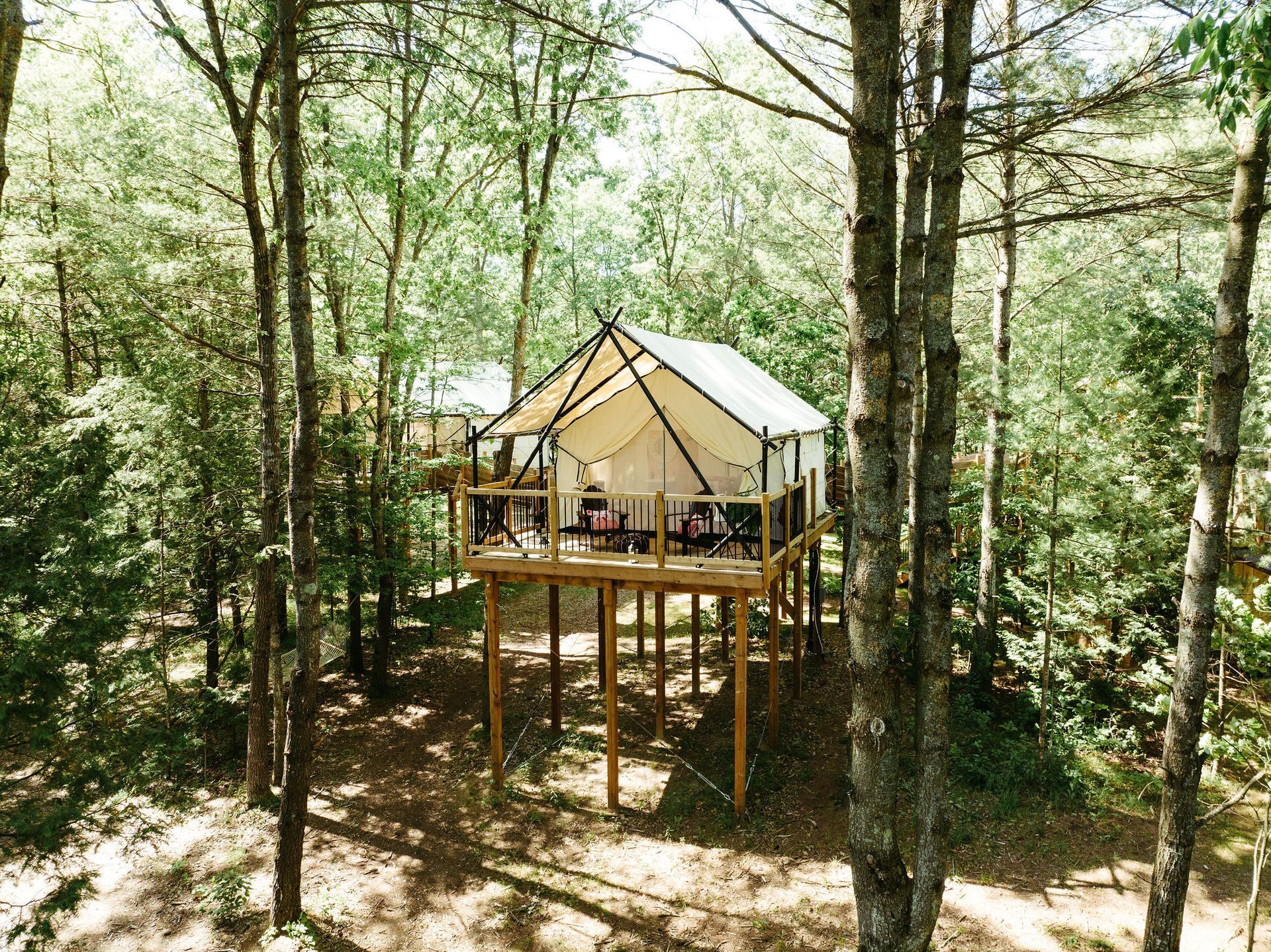 Canvas tent on stilts in a forest setting; wooden deck with furniture.