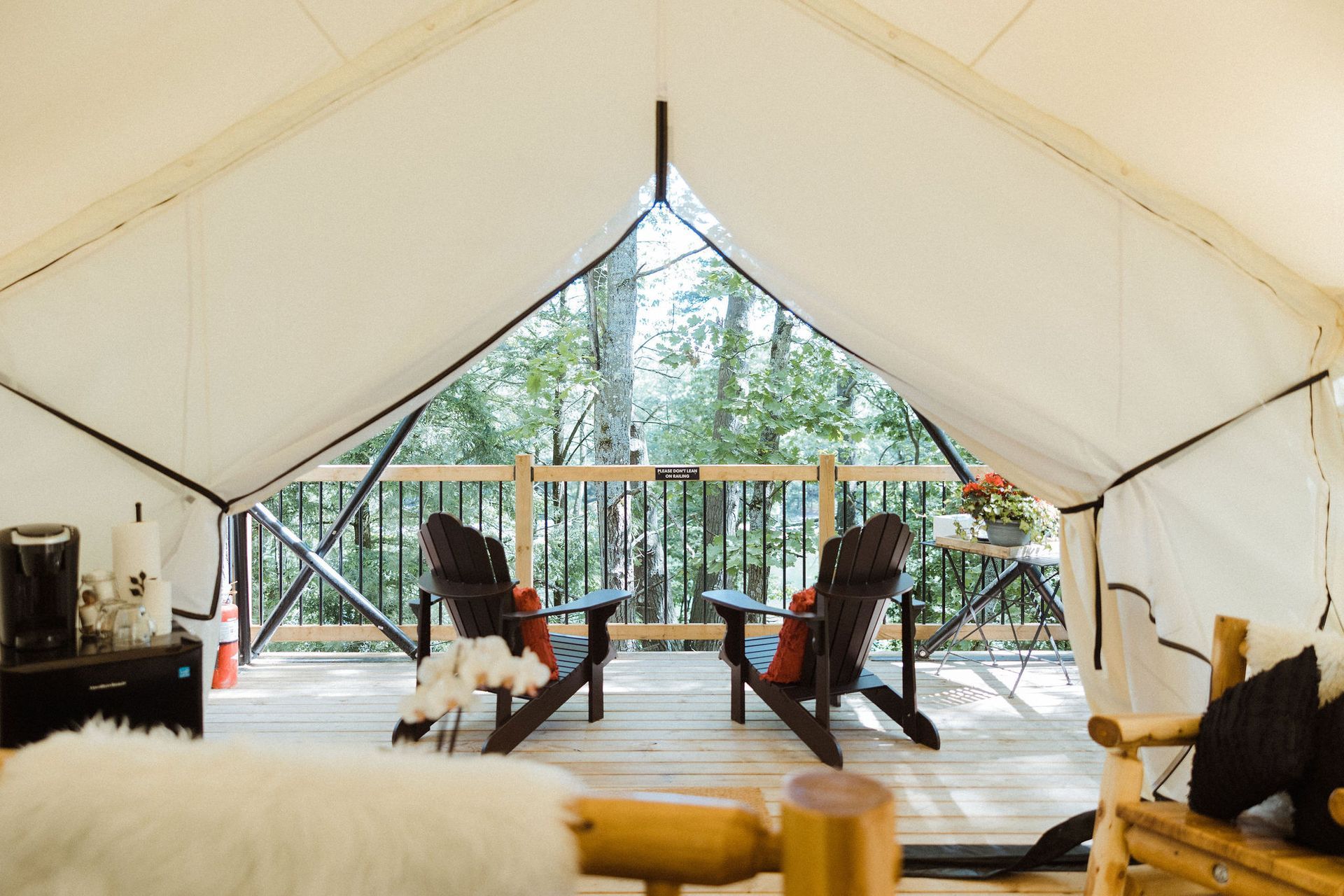 Interior of a canvas tent with two Adirondack chairs on a deck overlooking trees; a coffee maker is visible.