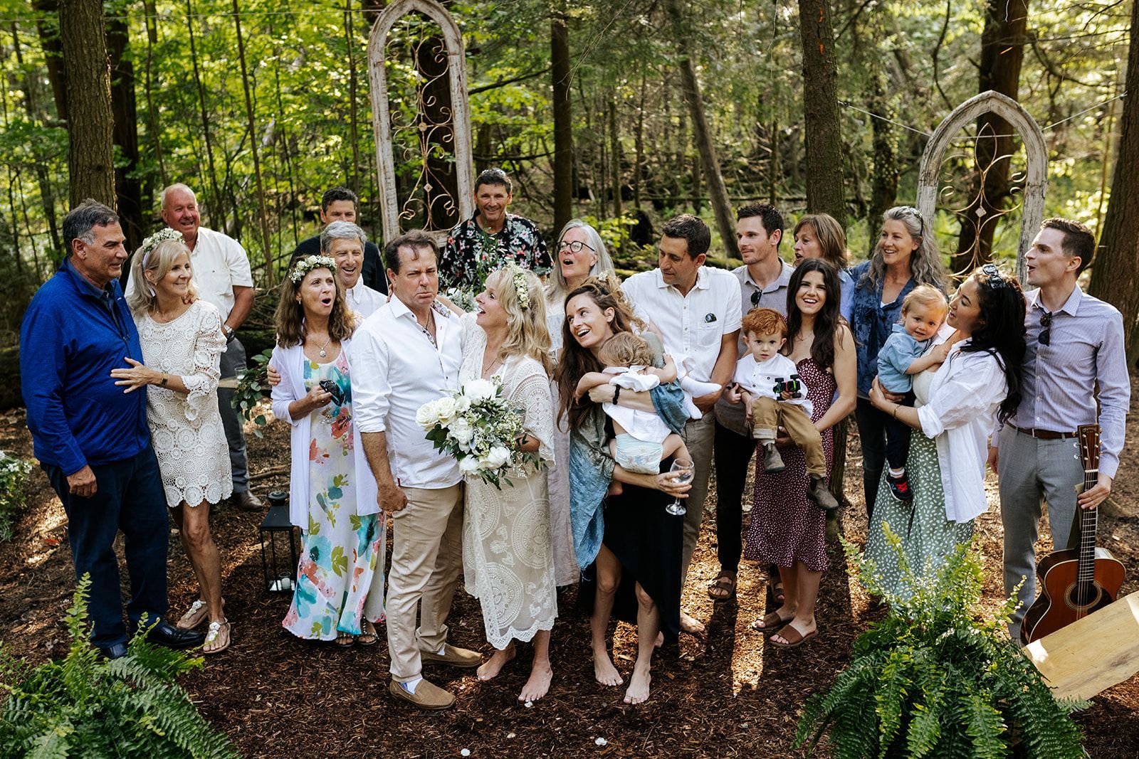 Wedding group in a forest setting; bride and groom centered, surrounded by family and friends, smiles, two white arches.