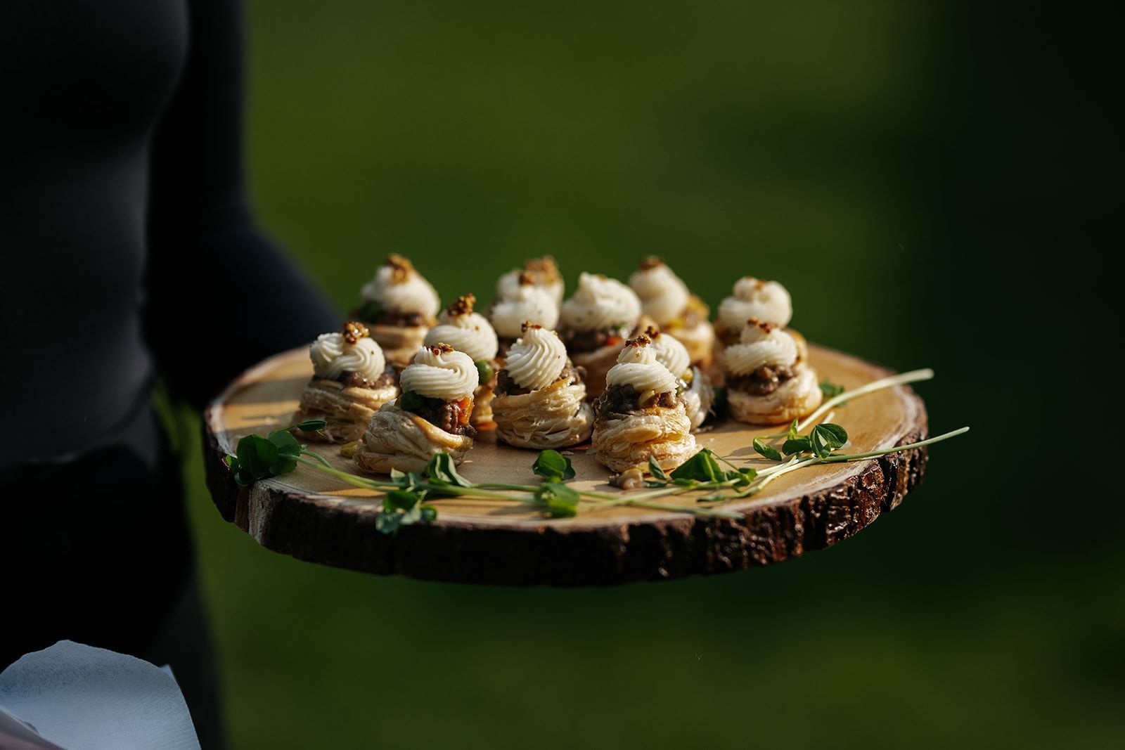 Server holding a wooden platter of appetizers outdoors; miniature food towers with garnishes.