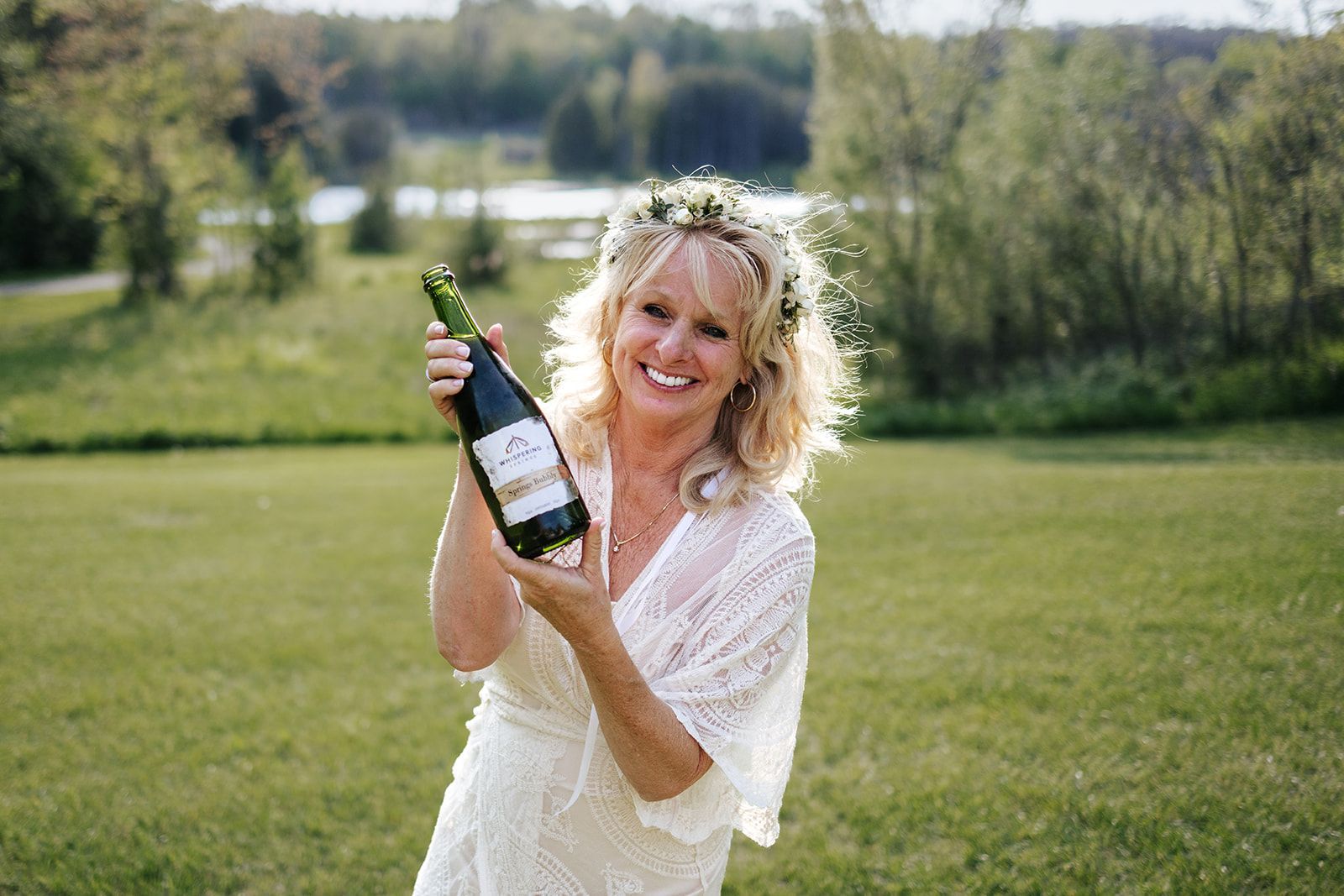 Woman in white dress and floral crown holding a champagne bottle, smiling outdoors.