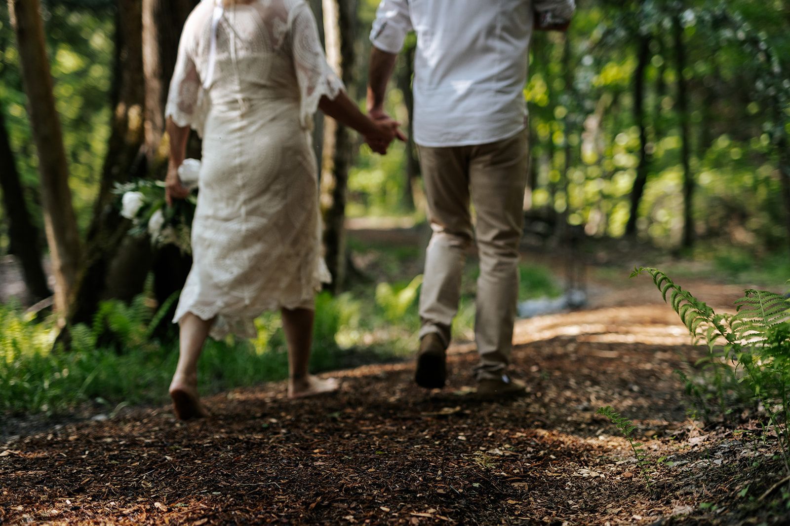 Couple holding hands, walking away on a forest path. Woman in white dress, man in tan pants. Green trees surround.