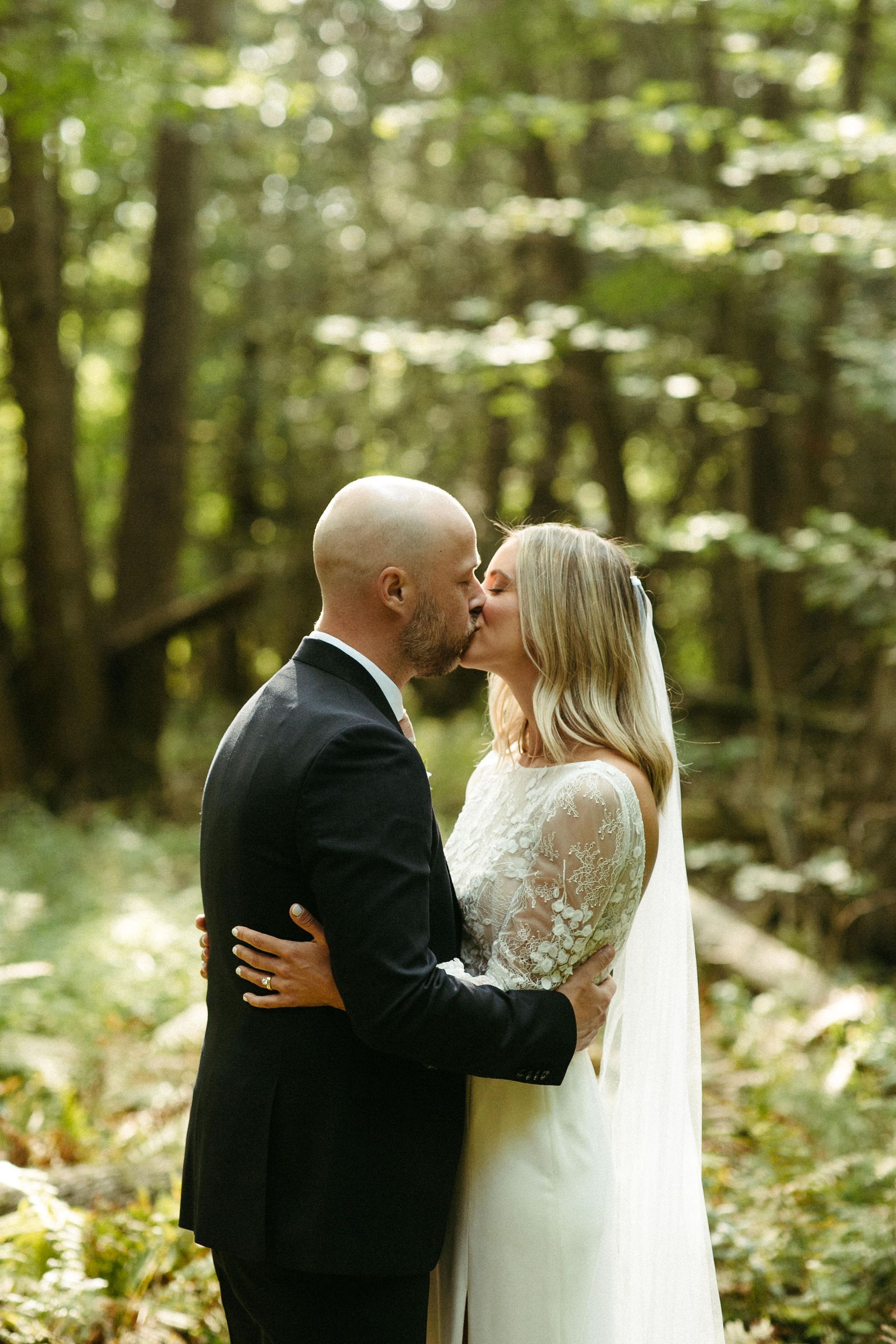 Couple kissing in a forest; the woman wears a white wedding dress with lace sleeves and veil; the man wears a black suit.