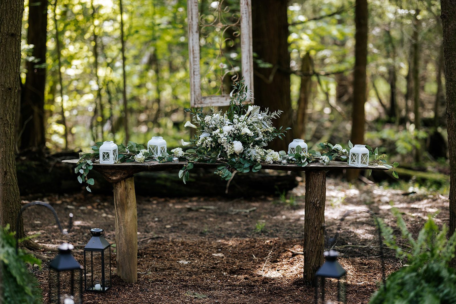 A woodland wedding altar. Wooden table with floral arrangement, lanterns, and a framed mirror.