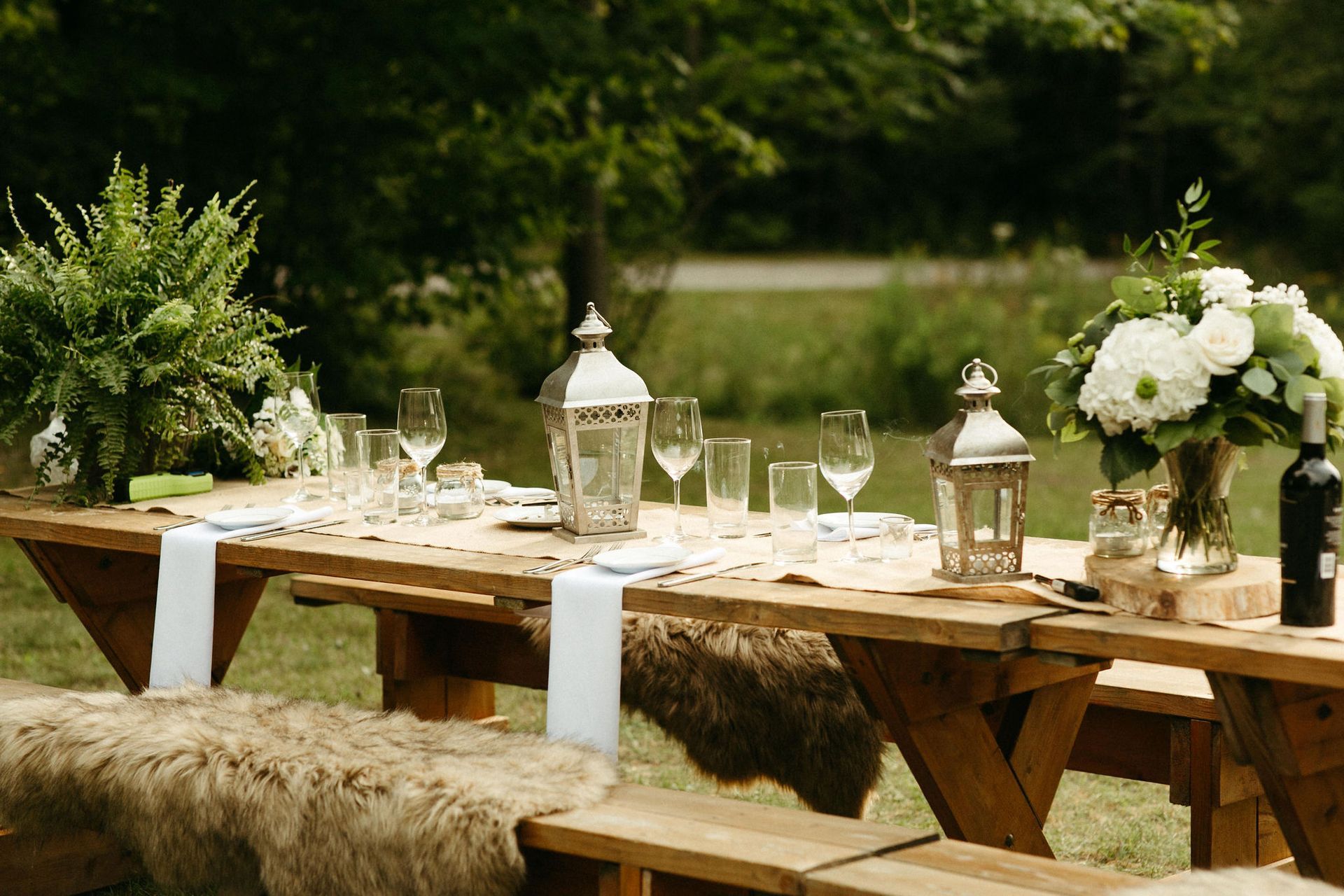 Outdoor table setting with lanterns, flowers, and glassware; picnic benches with fur throws.