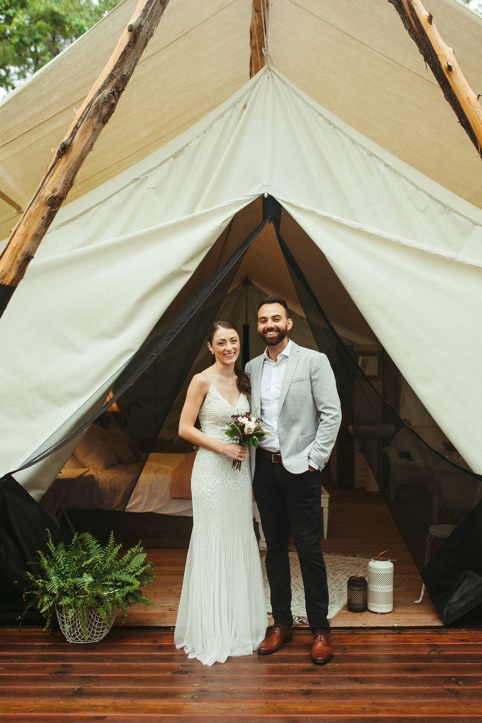 Bride and groom smiling in front of a white tent, holding flowers, on a wooden deck.