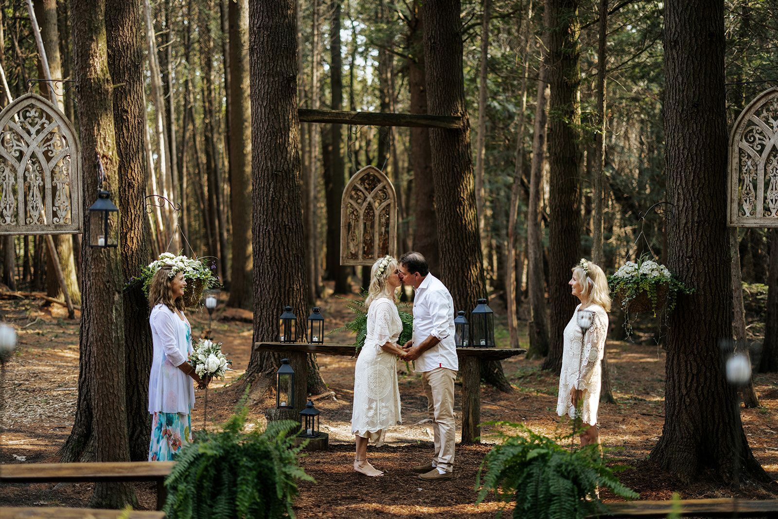 Wedding ceremony in a forest clearing: couple kissing, officiant, two guests, decorative arches, and ferns.