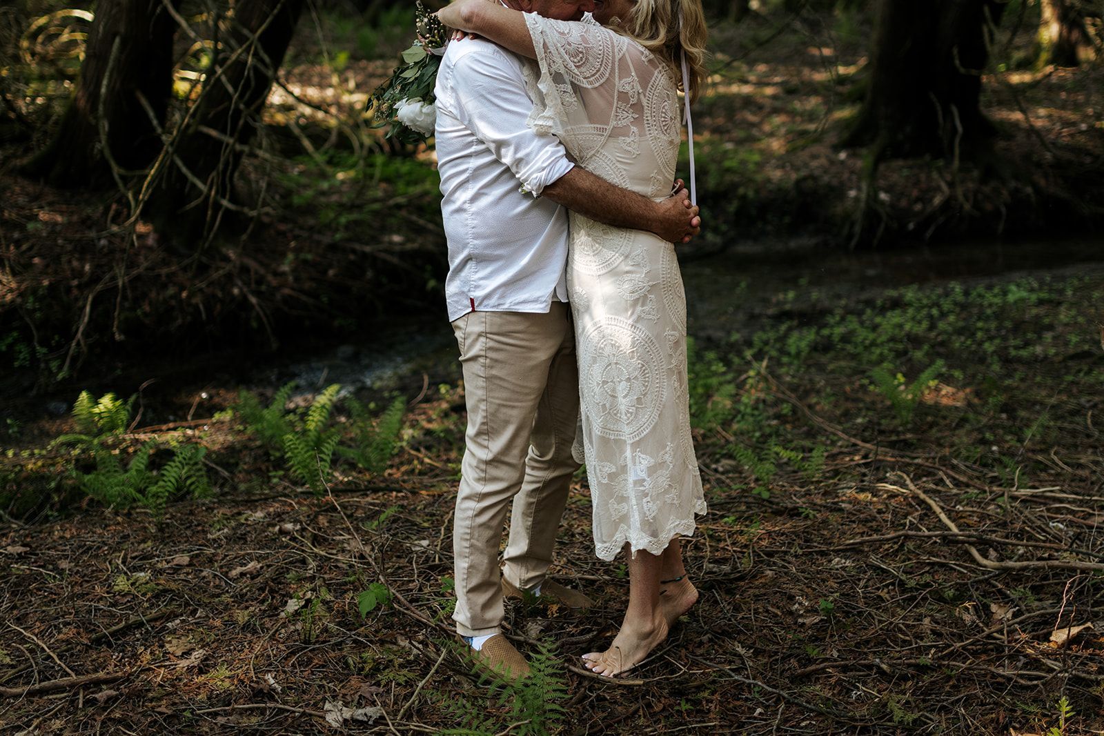 Couple hugging in a forest setting; the woman is barefoot, wearing a lace dress.
