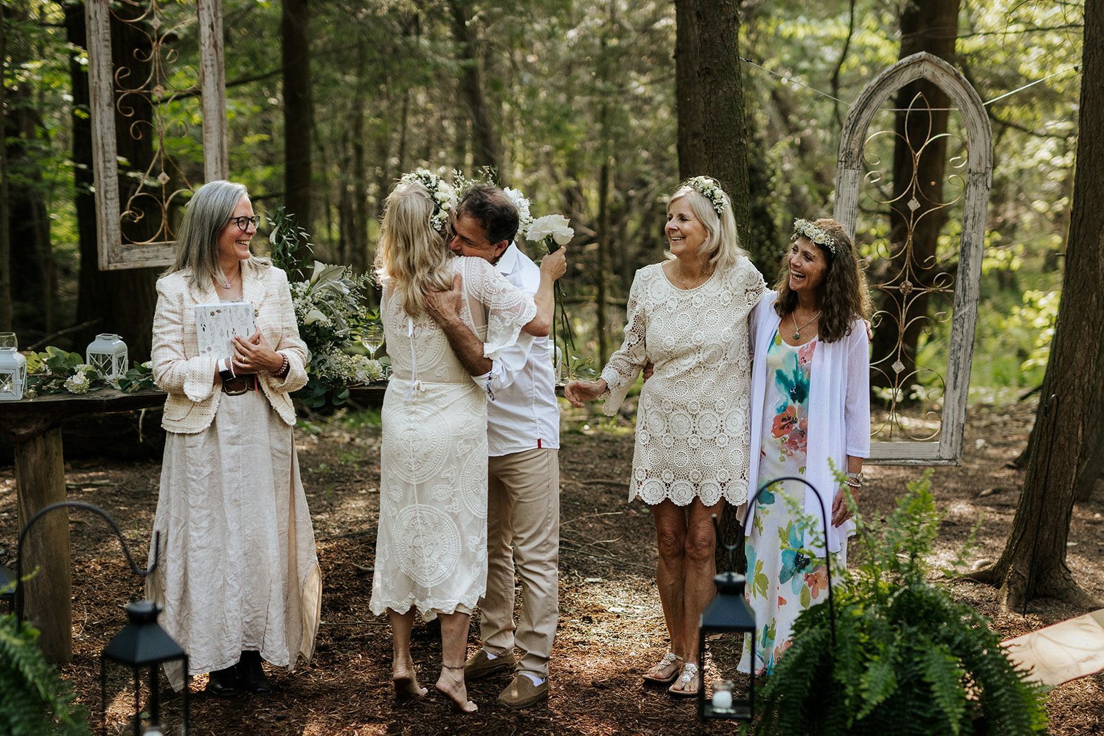 Wedding in a forest: couple embraces; guests smile. Floral decor, sunlight, trees.