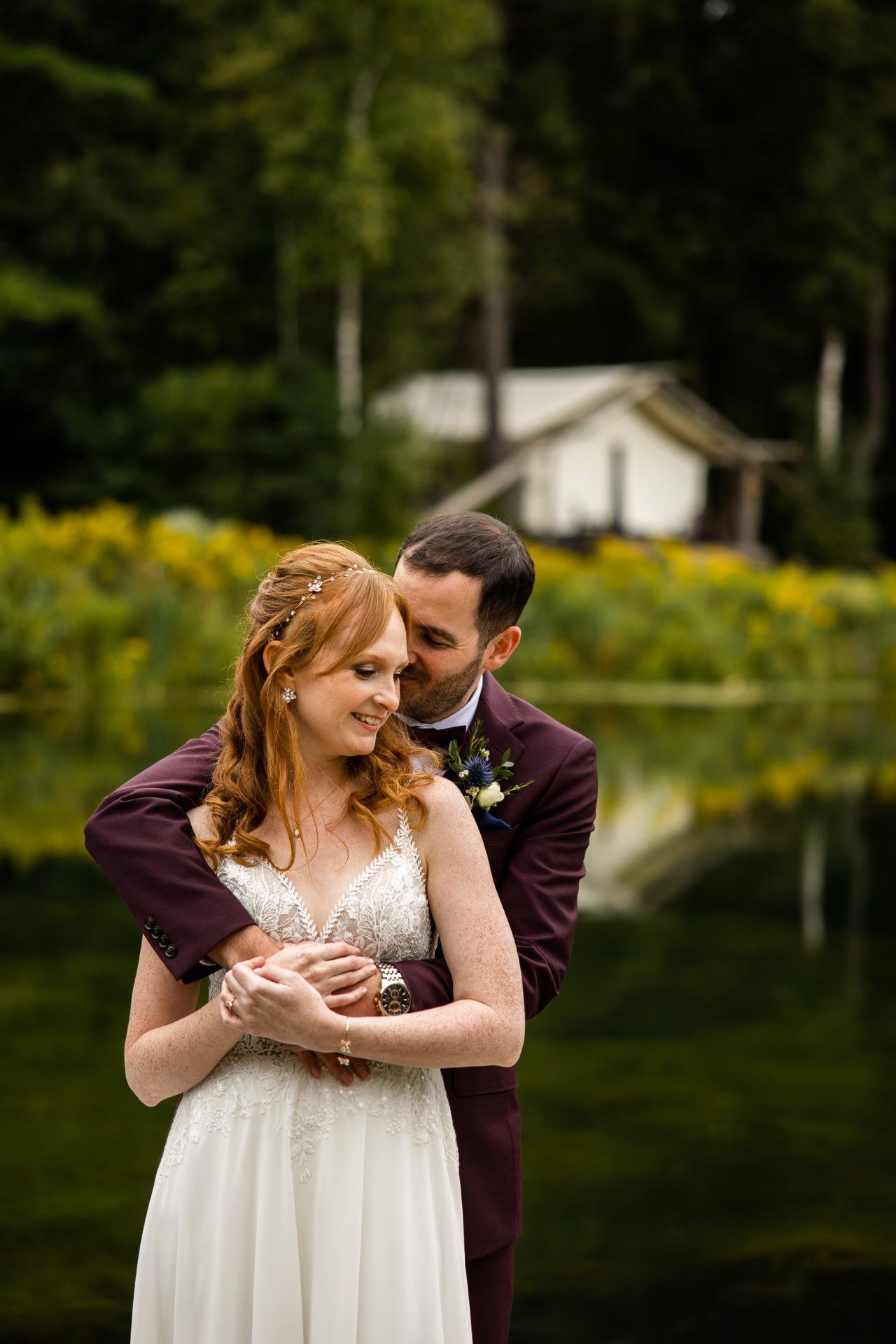 Newlyweds embrace near a lake; the man in a burgundy suit, the woman in a white dress, with a cabin in the background.