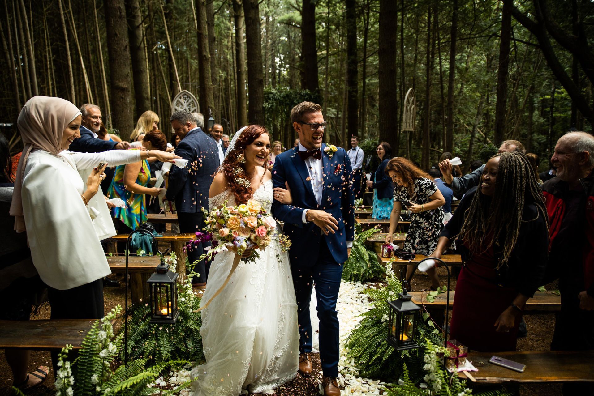 Newlyweds walk down aisle as guests toss confetti, outdoor forest wedding.