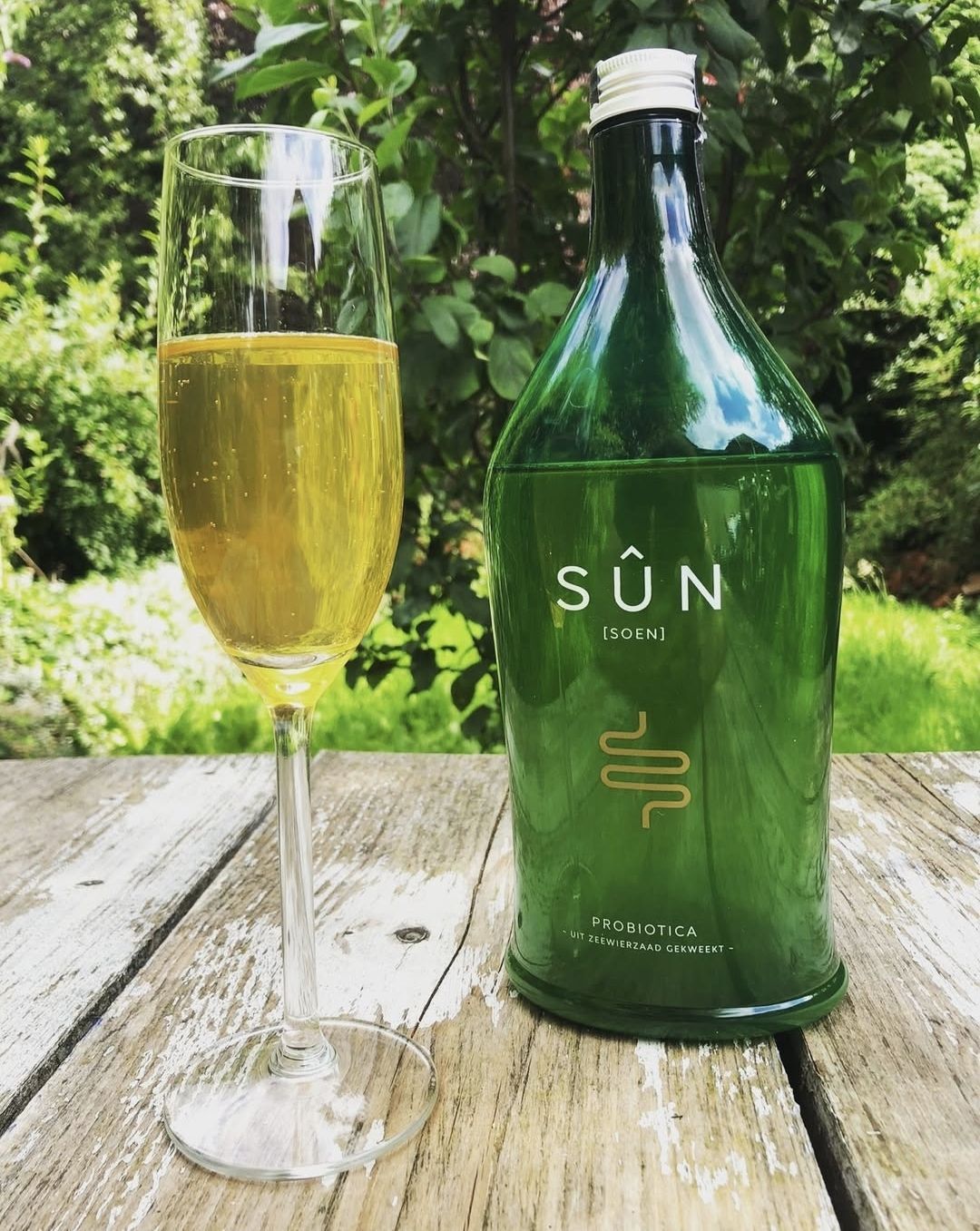 Een glas mousserende wijn naast een groene SÛN-fles op een verweerde houten tafel buiten.