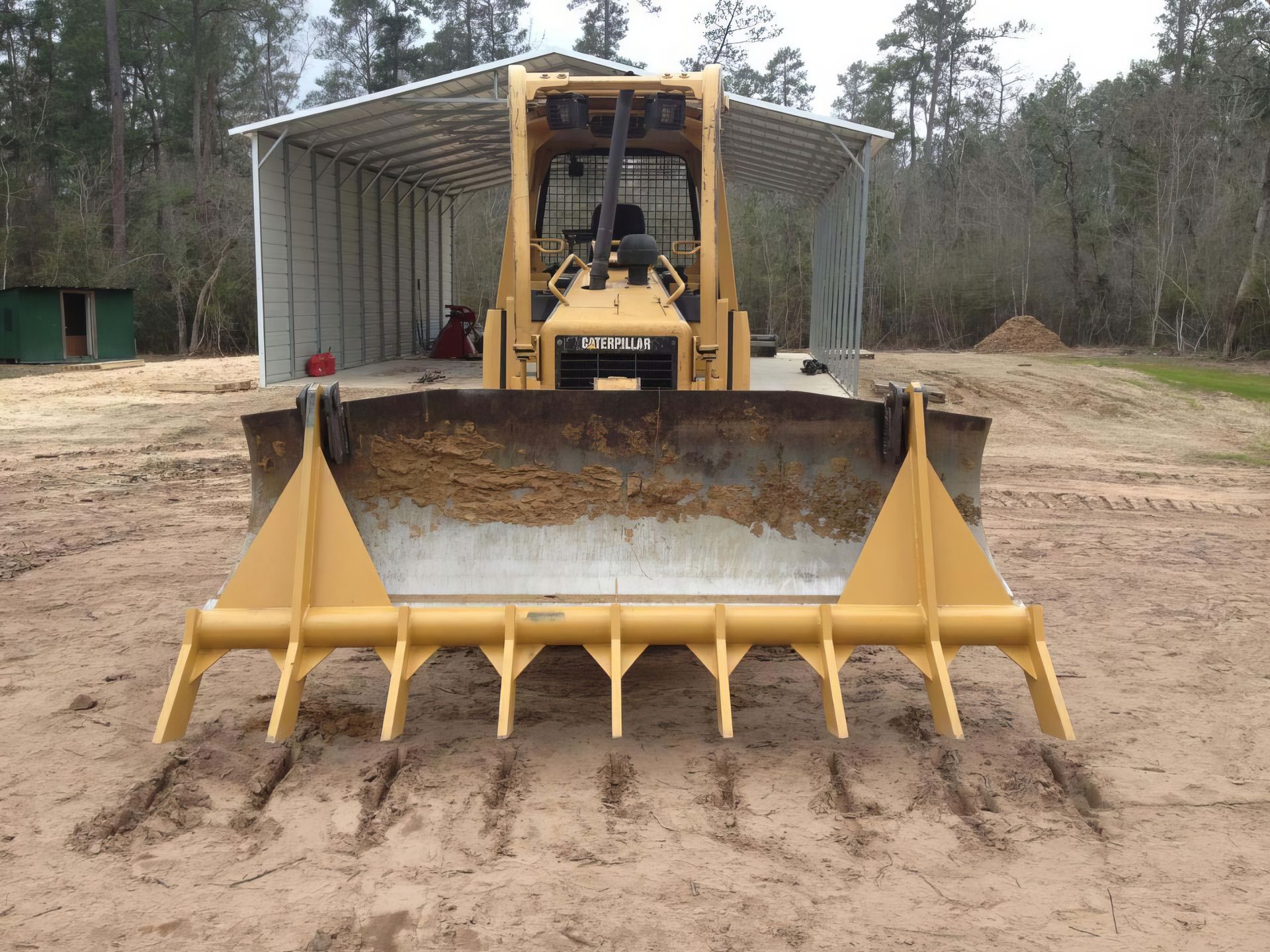 Yellow bulldozer with a front blade on a dirt road, facing an open shipping container in a wooded area
