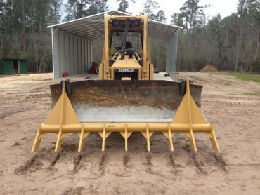 Yellow bulldozer with a front blade on a dirt road, facing an open shipping container in a wooded area