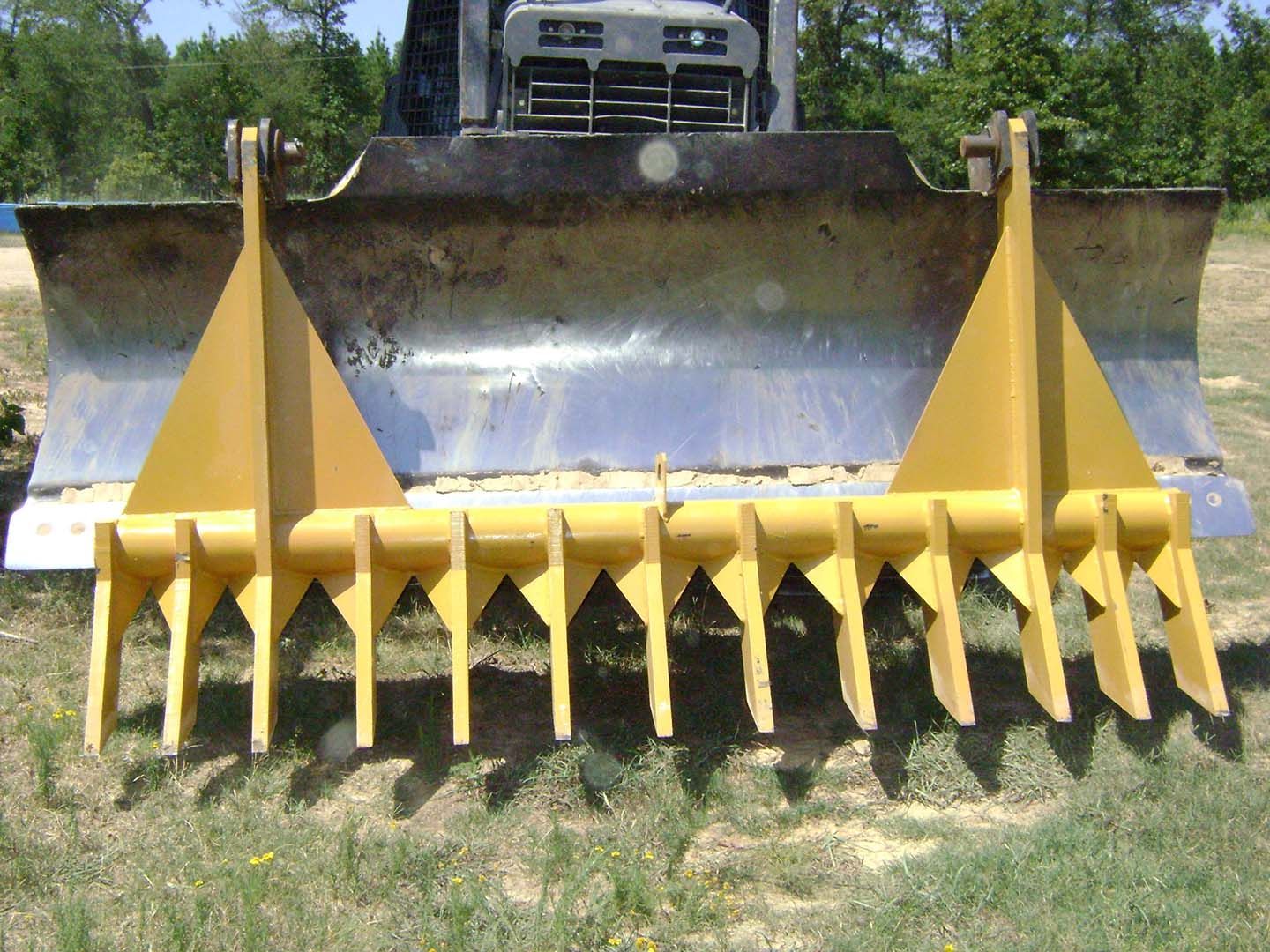 Yellow bulldozer snow plow attachment with sharp tines parked on grass in front of a truck