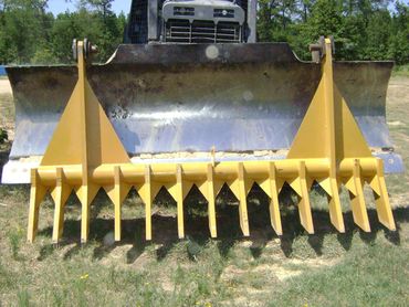 Yellow bulldozer snow plow attachment with sharp tines parked on grass in front of a truck