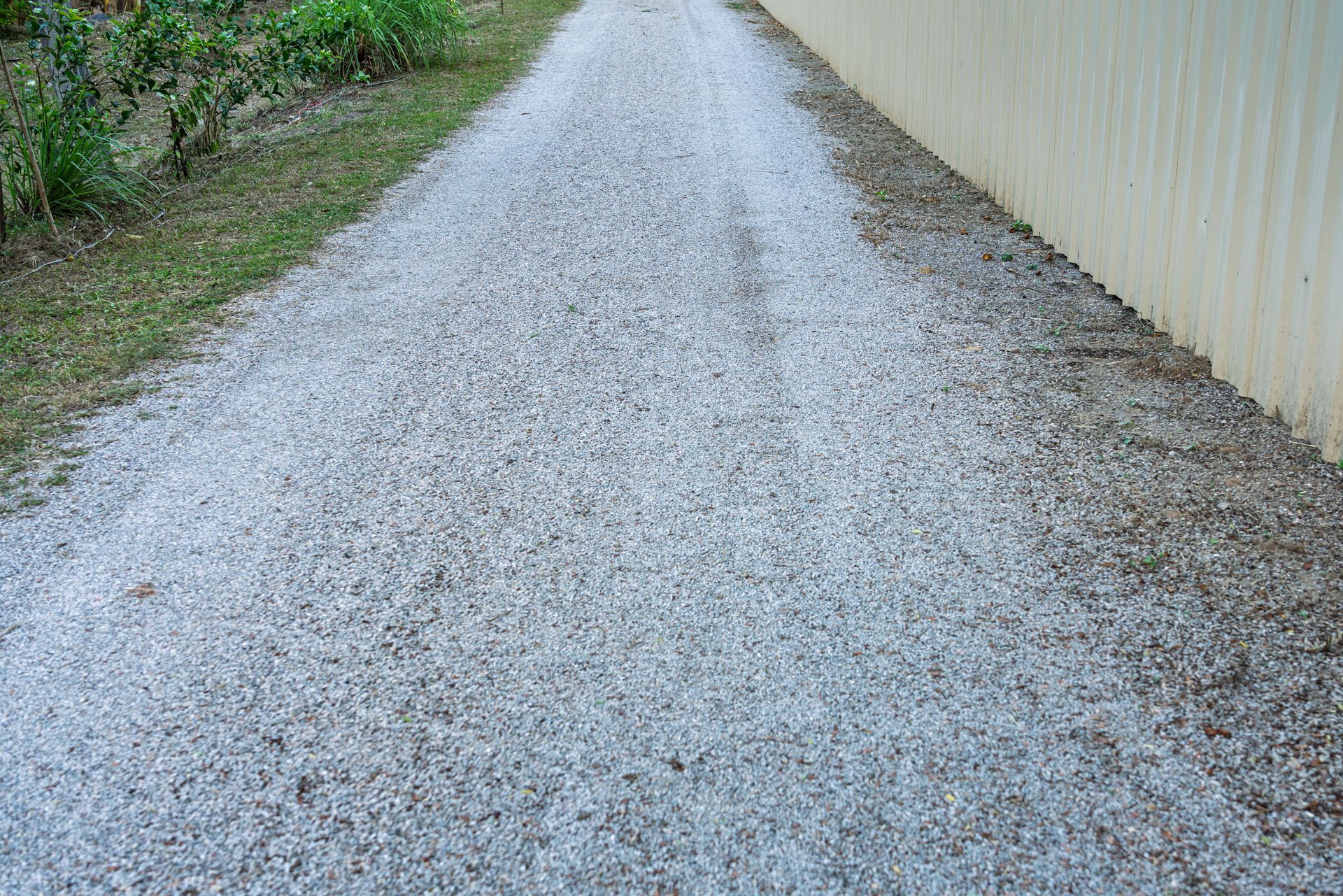 Gravel driveway beside a beige building, with greenery along the left edge.