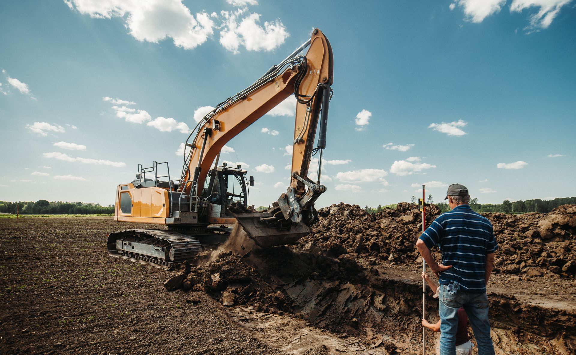 Excavator digging a dirt trench in a field while a man in a blue striped shirt stands nearby under a cloudy sky