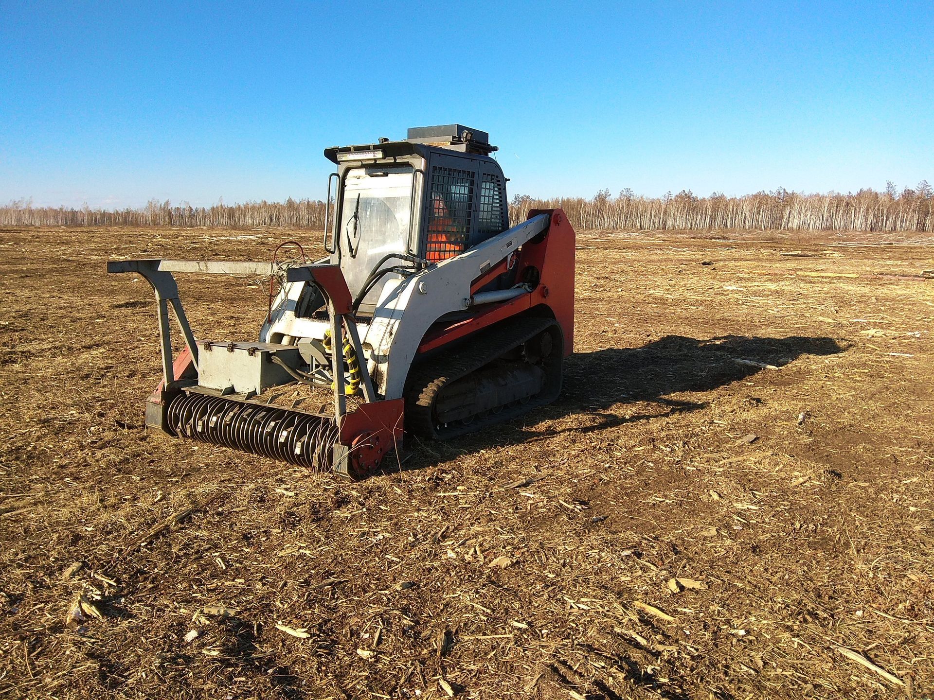 Tracked harvester in a harvested field under a clear sky, clearing crop residue with a front attachment