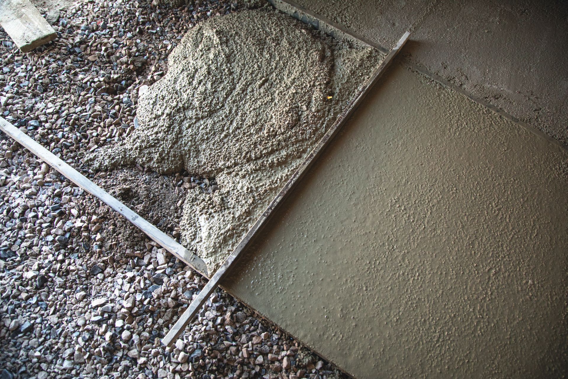 Fresh cement being spread beside a gravel surface, with a trowel visible in the corner
