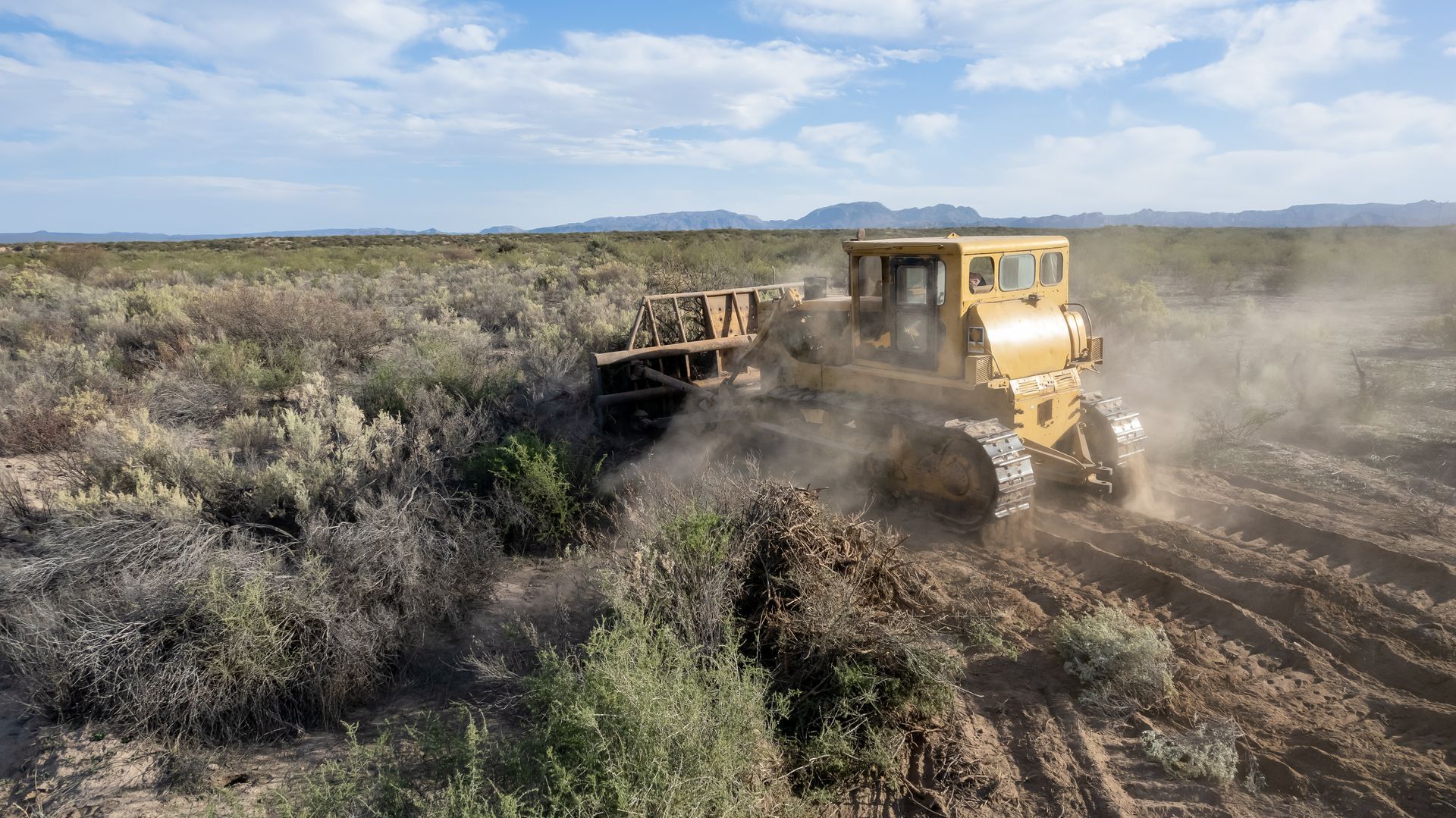 Yellow bulldozer grading a dusty dirt road through scrubland under a cloudy sky.