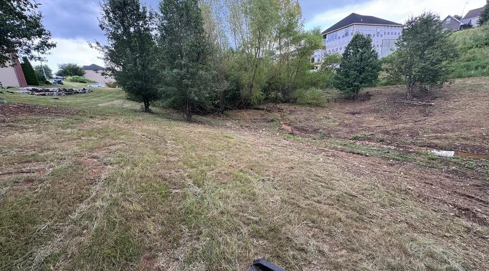 Grassy vacant lot with a dirt path, trees, and a house in the background