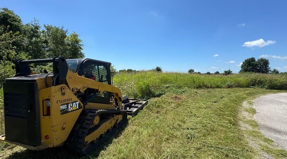 Yellow skid steer clearing grass beside a paved road under a blue sky