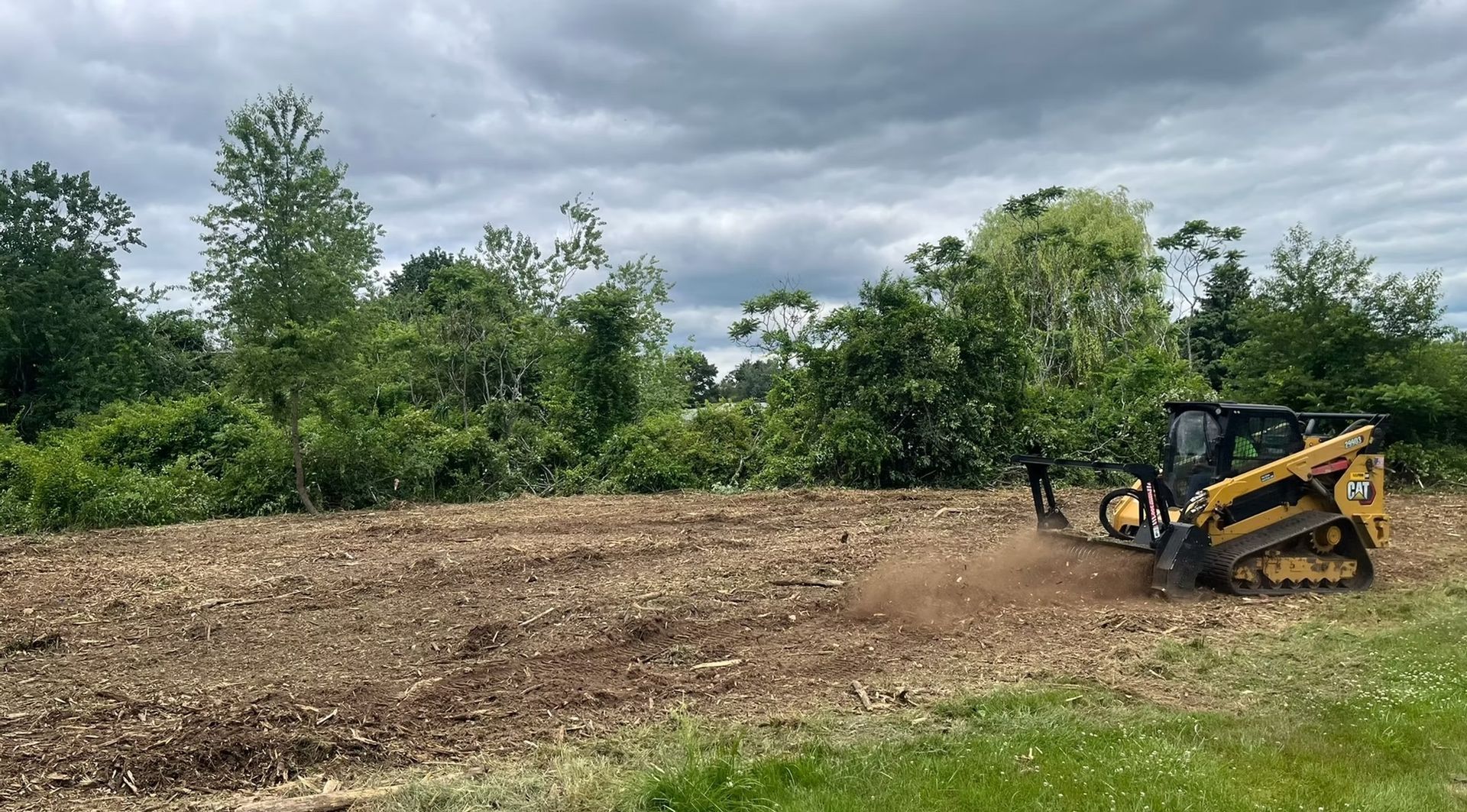Small skid steer clearing a dirt field with trees and cloudy sky in the background