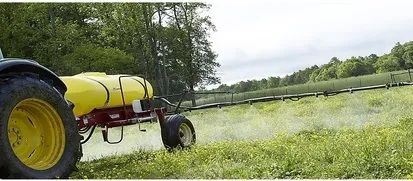 Yellow agricultural sprayer spraying crop field with mist, viewed from the side under a cloudy sky