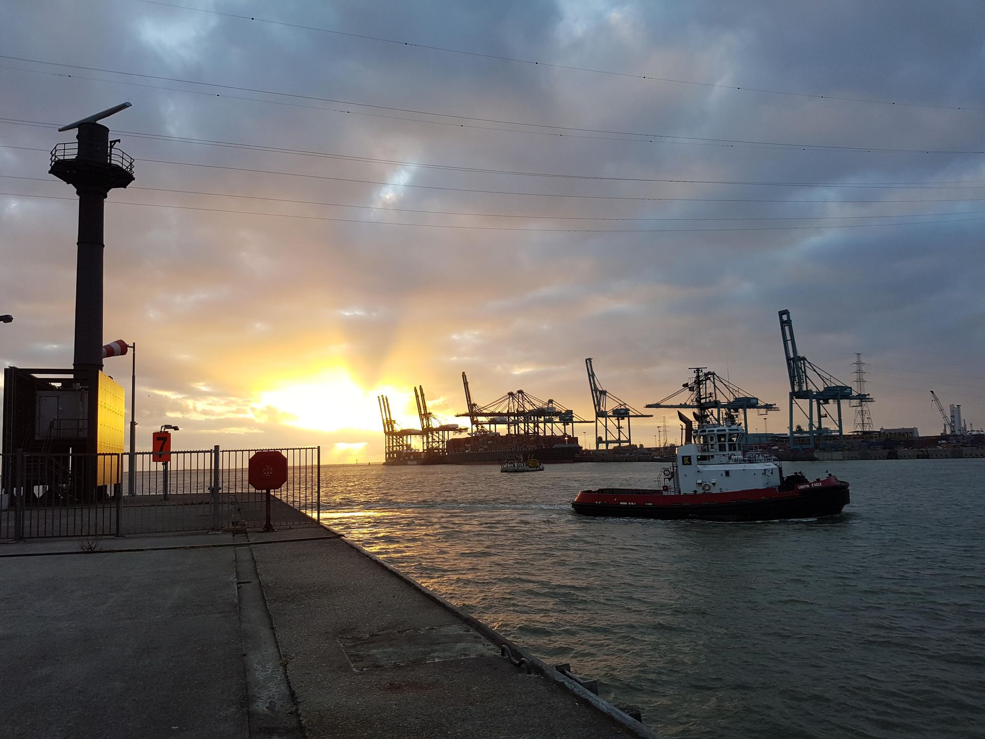 Lever de soleil sur un port avec des grues, un bateau et un phare. Ciel nuageux.