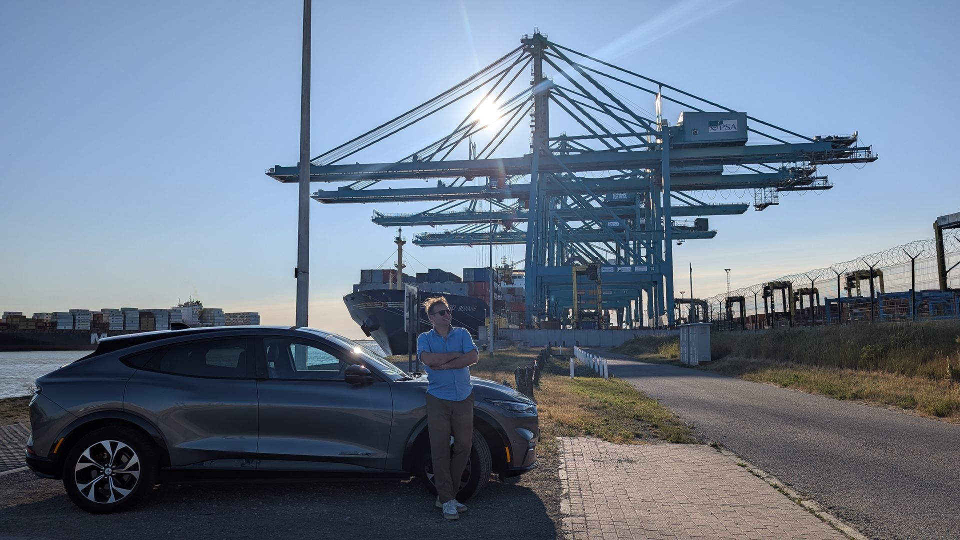 Man standing by a gray electric car, arms crossed, with shipping cranes and a ship in the background. Sunny, outdoor setting.