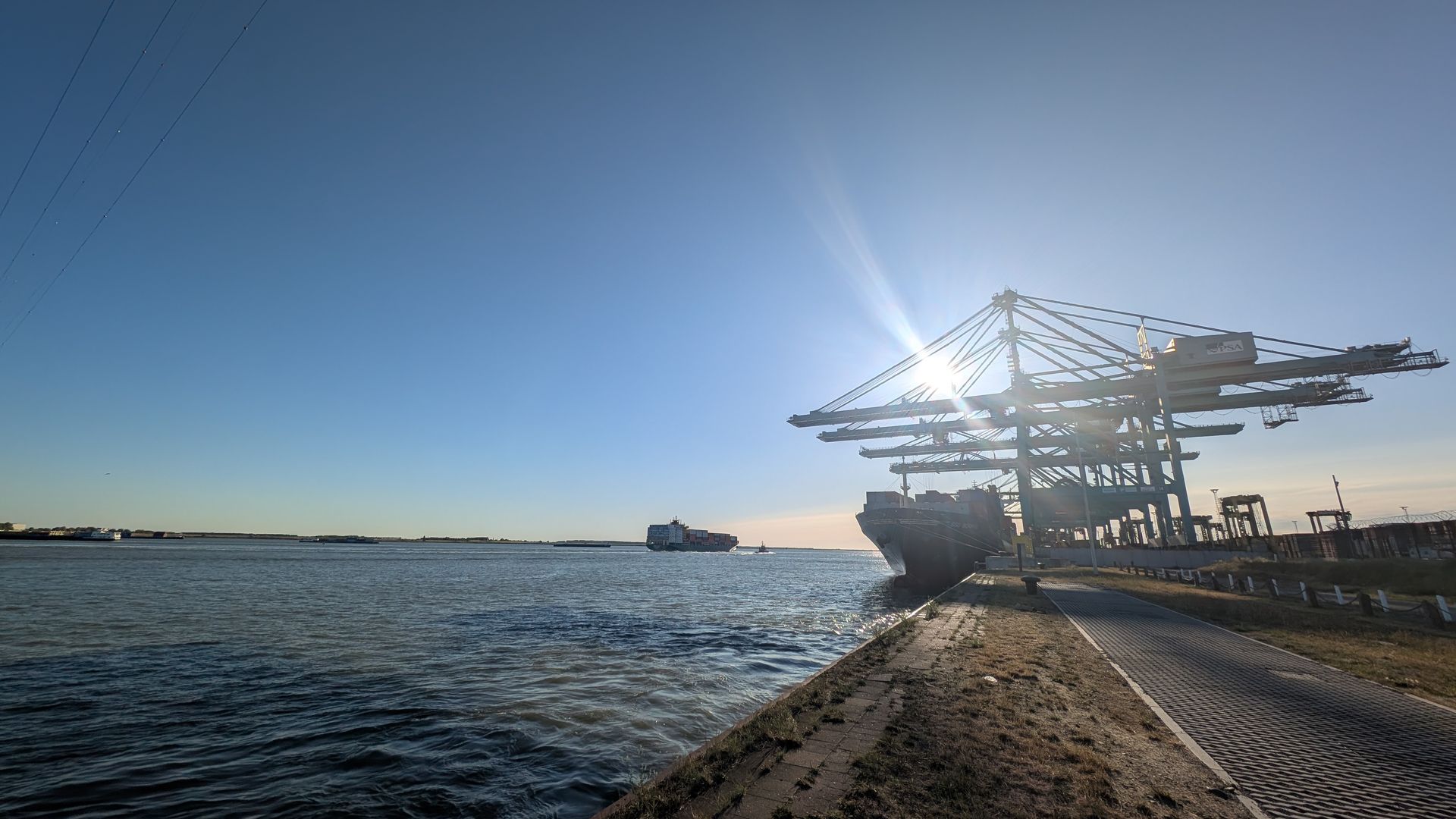 Container ship at a sunny port, silhouetted by the sun. A crane looms over the ship.