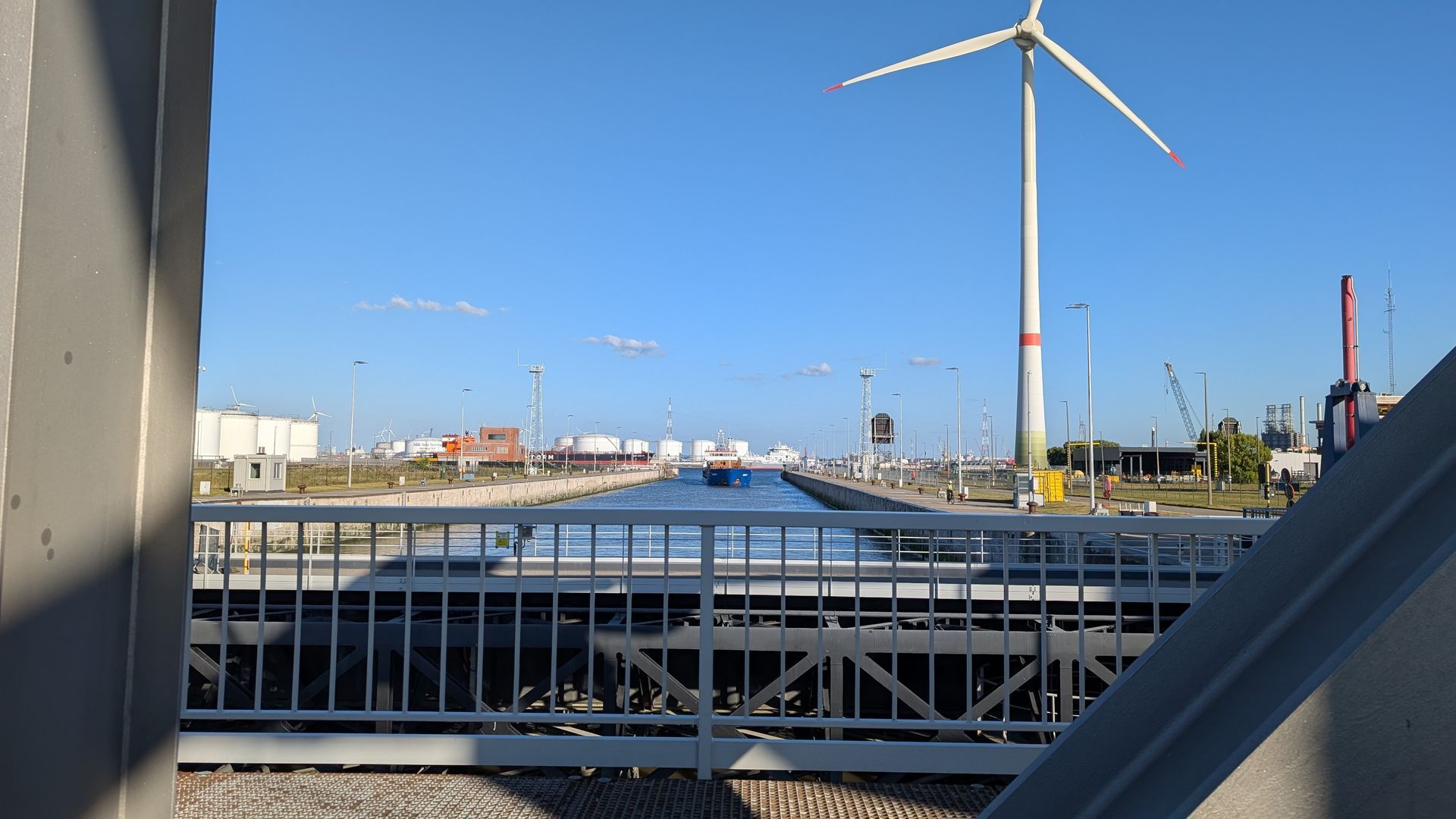 View of a canal lock with a wind turbine, blue sky, and industrial buildings in the background.
