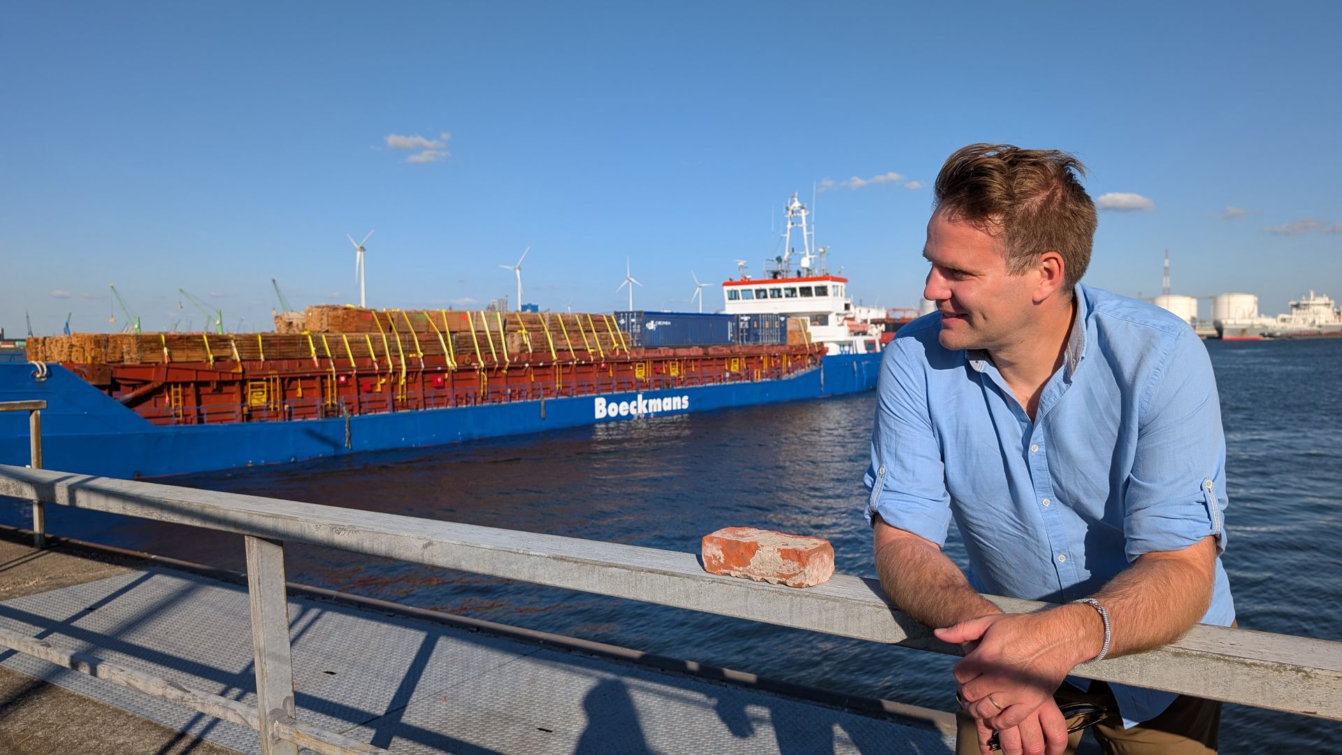 Man leaning on a railing, looking at a blue cargo ship at a harbor on a sunny day.