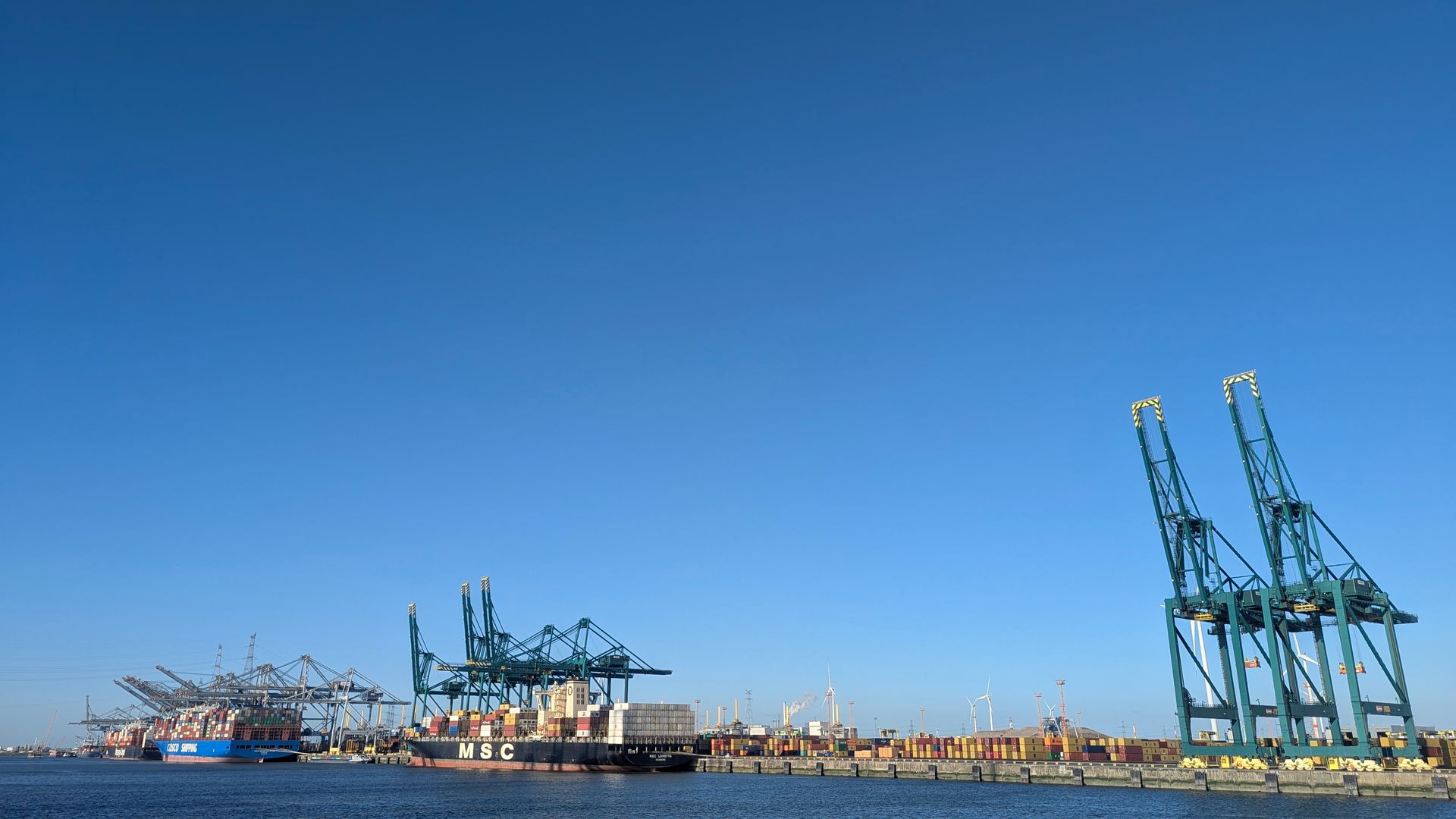 Blue sky over a harbor with large teal cranes and cargo ships, and industrial infrastructure.