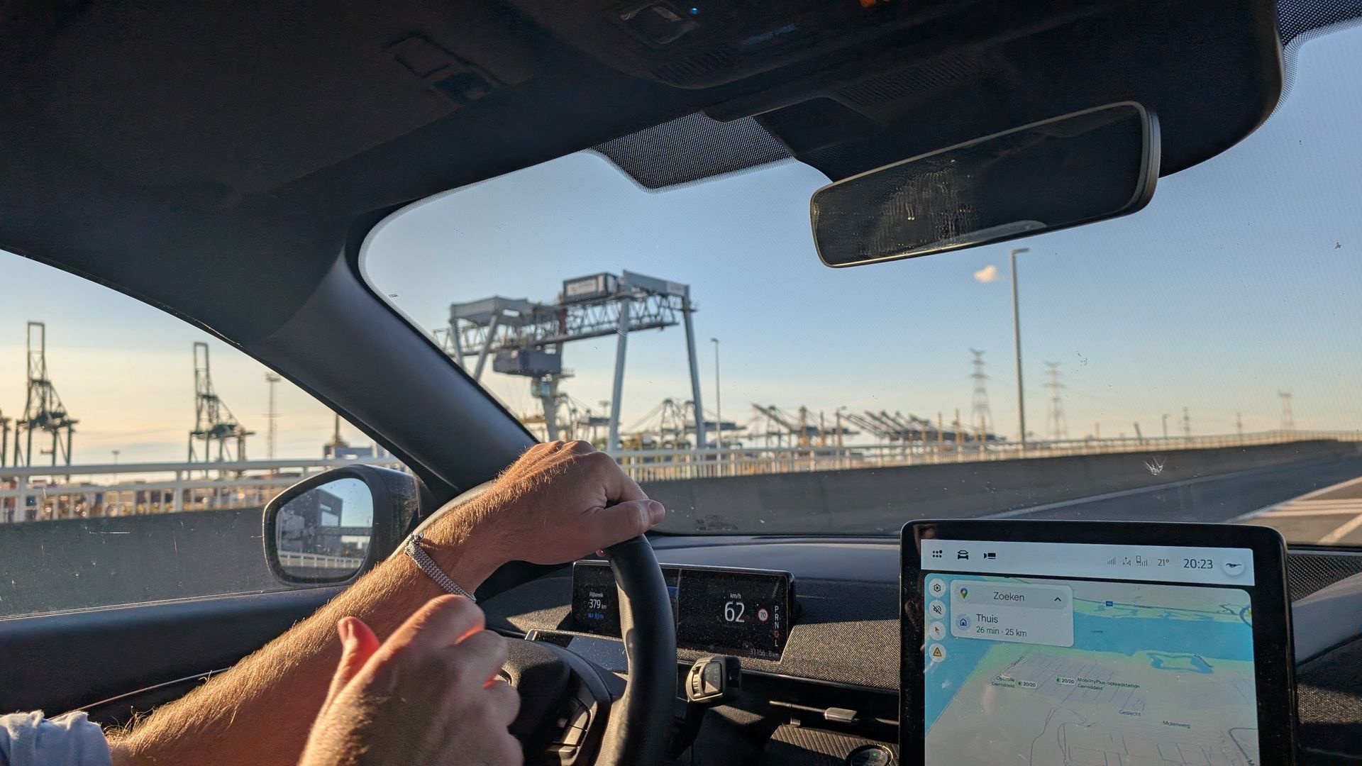 Person driving a car on a bridge, hands on the steering wheel, port cranes visible in the background.