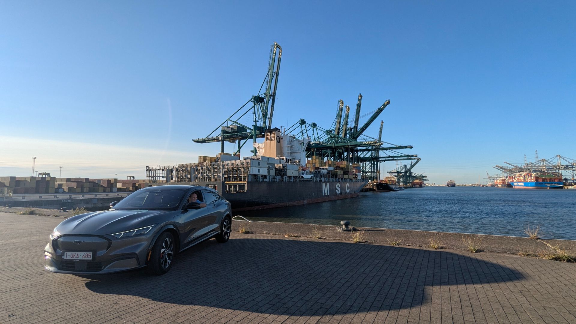 A gray Ford Mustang Mach-E parked at a port, cargo ship and cranes in the background under a blue sky.
