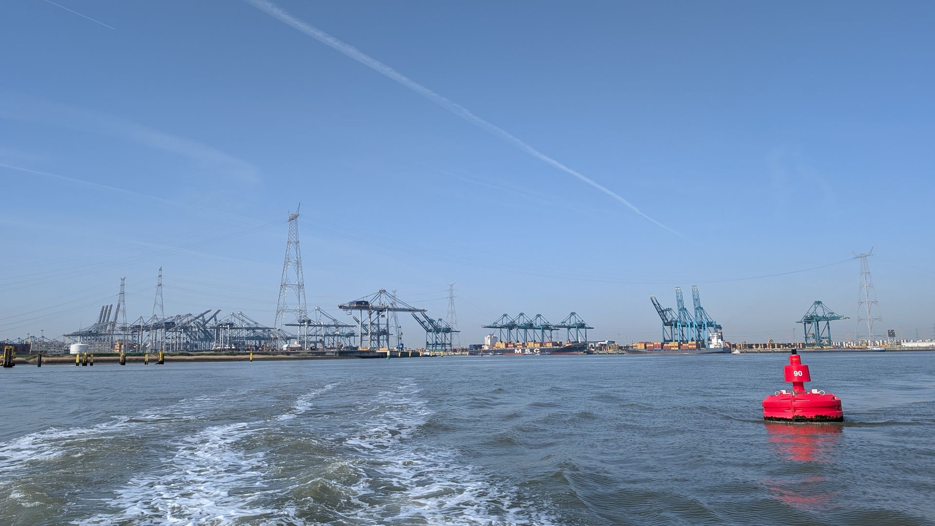 Harbor with cranes and ships under a blue sky, seen from a boat leaving a white wake, with a red buoy in front