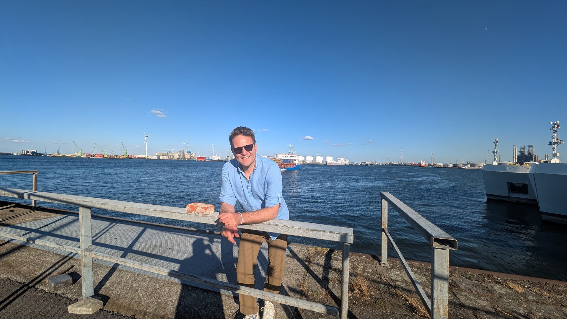 Man in sunglasses leans on a railing, overlooking water with a harbor and blue sky in the background.