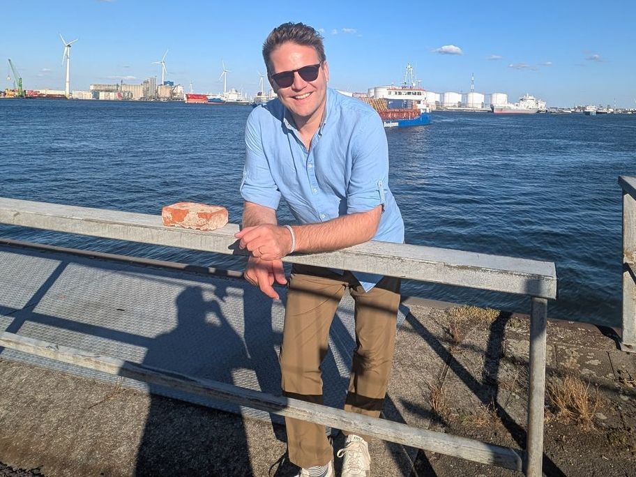 Homme portant des lunettes de soleil, appuyé sur une balustrade en métal, souriant au bord de l'eau avec des navires 