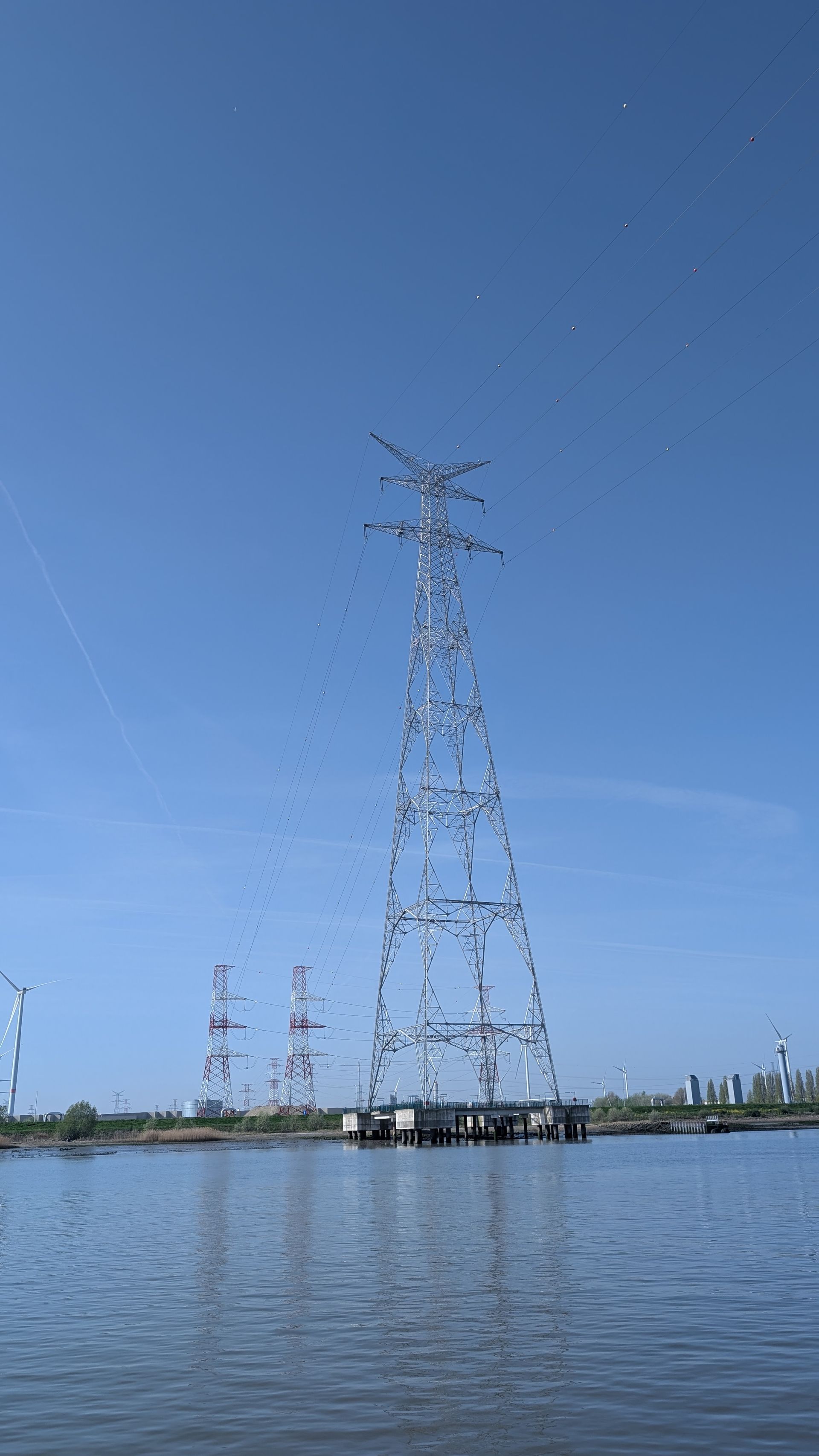 Tall power transmission tower rising from water under a clear blue sky, with smaller towers in the distance