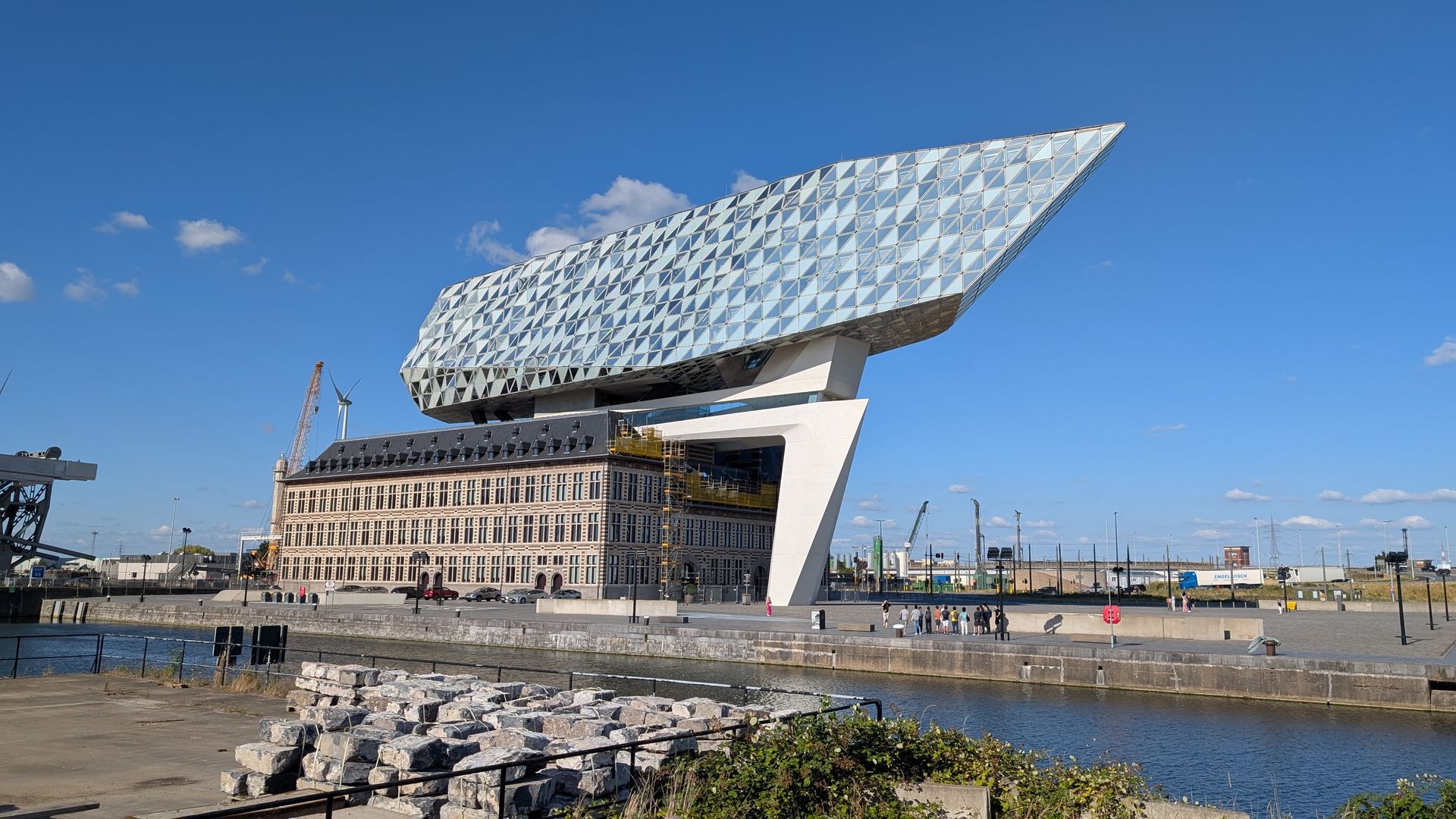 Modern building in Antwerp, Belgium, with a glass facade atop an old brick structure, next to a body of water.