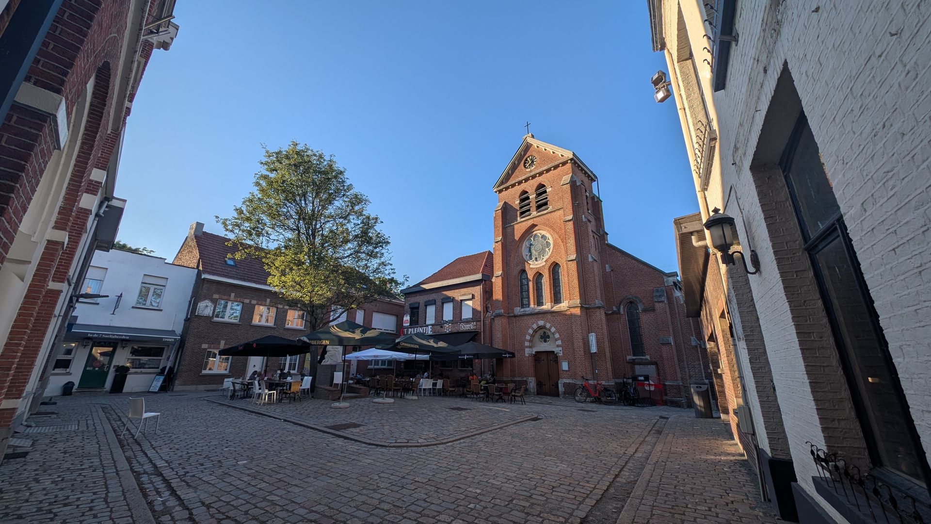 Brick church with a clock tower in a cobblestone square, flanked by buildings. Clear blue sky overhead.