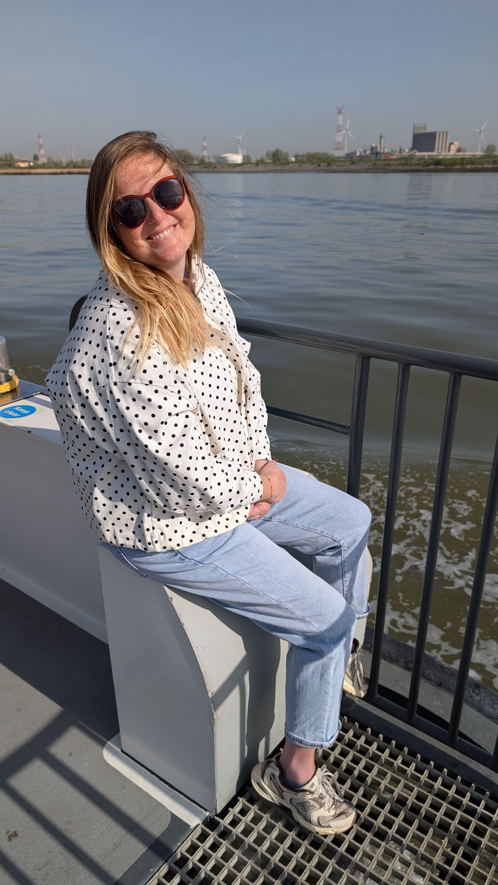Person sitting on a railing by the water, wearing sunglasses, a white patterned shirt, and jeans.
