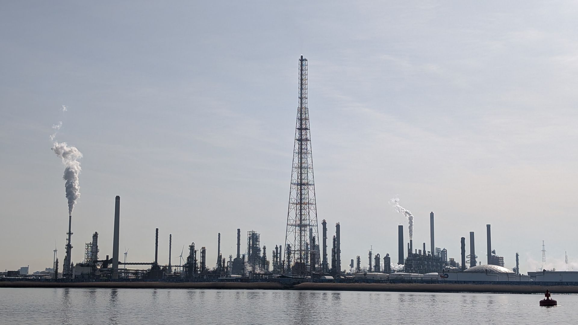 Industrial refinery skyline with tall smokestack and towers, emitting smoke beside calm water.